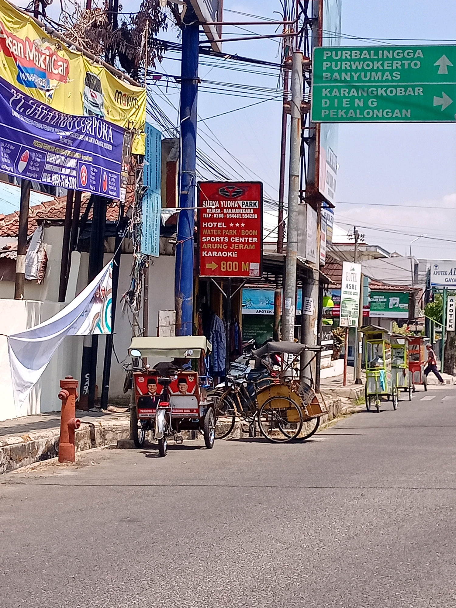The image shows a street scene in Banjarnegara, Indonesia, likely in the downtown area, based on the presence of various commercial signs and the density of the urban environment. In the foreground, the asphalt road is visible with lane markings. To the left, a bright orange fire hydrant stands near a raised sidewalk. Several motorized rickshaws, known as "becak motor," are parked or in motion. One becak motor in the mid-left foreground has images of two men in formal attire on its side, likely political figures. Further down the street, other becak motors and street vendors' carts are visible.
Overhead, a complex arrangement of utility poles and wires crisscrosses the sky, supporting numerous signs. These signs include advertisements for "Maks Car" and "PT. SATUTAMA CORPORA," a blue banner with text and logos, and a red sign for "SURYA YUDHA PARK" which also lists "HOTEL," "WATER PARK," "BOOM," "SPORTS CENTER," "ARUNG JERAM," and "KARAOKE," indicating a recreational facility. A directional signpost in green is also prominent, with arrows and text indicating destinations like "PURBALINGGA PURWOKERTO BANYUMAS" and "KARANGKOBAR DIENG PEKALONGAN."
A white banner, possibly a flag or promotional material, is draped over a pole on the left side of the image. The buildings lining the street are mostly two to three stories high, with tiled roofs visible. The overall atmosphere is that of a busy street on a bright, sunny day.