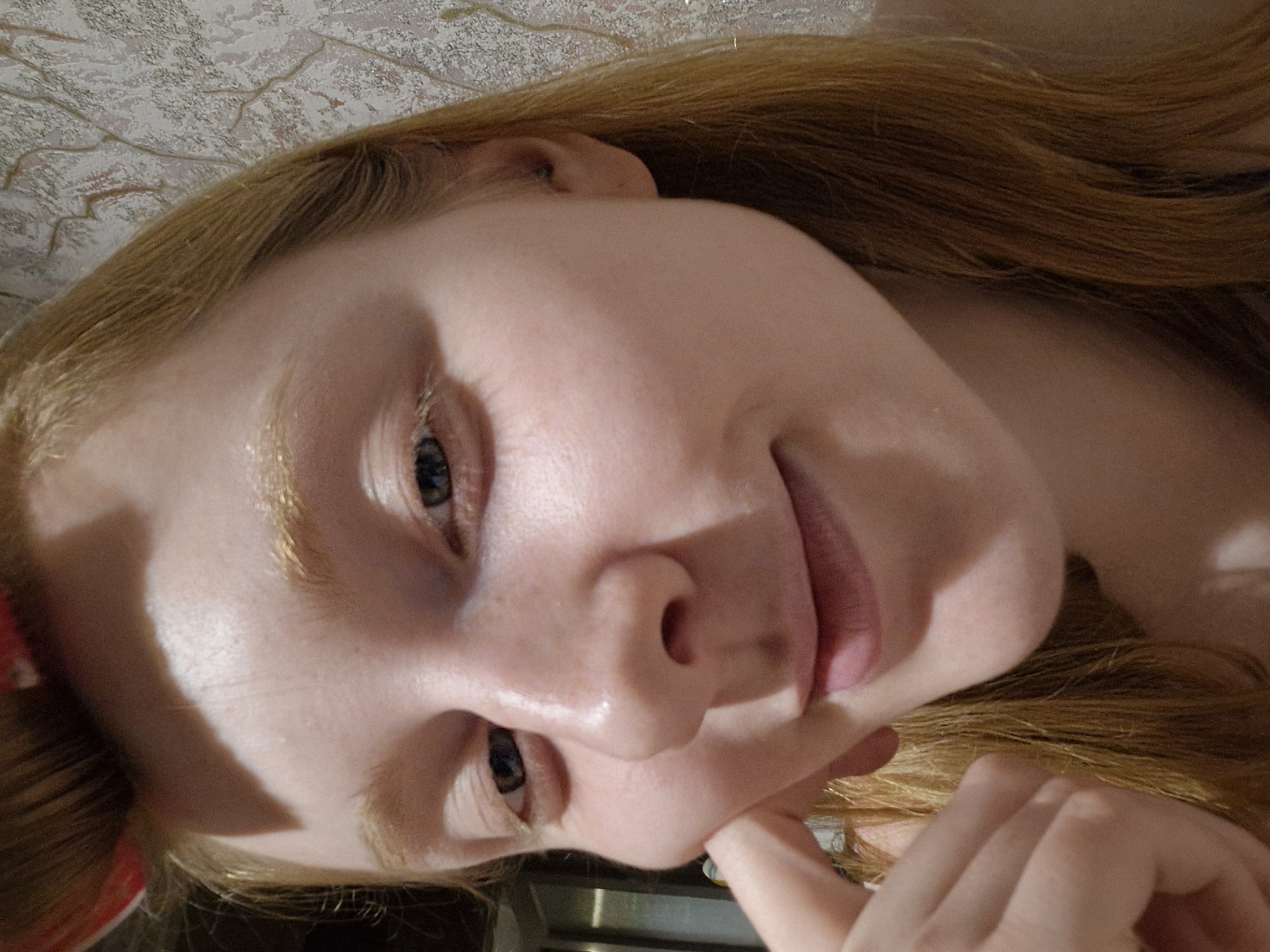 A fair-skinned person with light reddish-blonde hair is shown in a close-up, slightly tilted view. The person has blue eyes, a slightly upturned nose, and full lips. The person's hand is near the chin as if in a pensive pose. The background is blurry and appears to be a patterned surface, potentially a wall or bedding. The location of the image is not identifiable as Yekaterinburg, Russia.