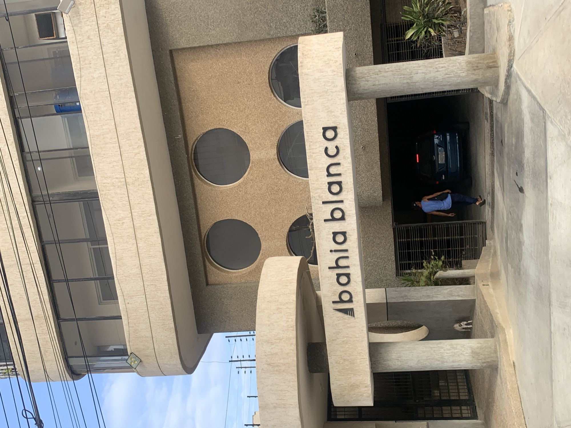 An exterior, daytime shot captures a modern, multi-story building in Porlamar, Venezuela. The building features textured beige and brown facades, with large rectangular windows on the upper floors and a series of circular windows on a lower section.
Prominently in the foreground, a tall, vertical sign structure displays the text "bahia blanca" in black, sans-serif font, preceded by a small triangular symbol. The sign and portions of the building's lower structure are made of a light, textured material.
At ground level, a covered entrance or garage area is visible, where a dark blue or black car is parked. A person wearing a blue shirt and dark pants is walking into this covered area, away from the viewer. Some utility lines are strung across the clear, bright blue sky with scattered clouds in the upper left of the frame. Small patches of green vegetation are visible near the building's base. A small, black and white dog is partially visible near the base of the sign. A square outdoor light fixture is attached to the building's facade. The scene depicts a quiet, ordinary moment at what appears to be a residential or mixed-use building.