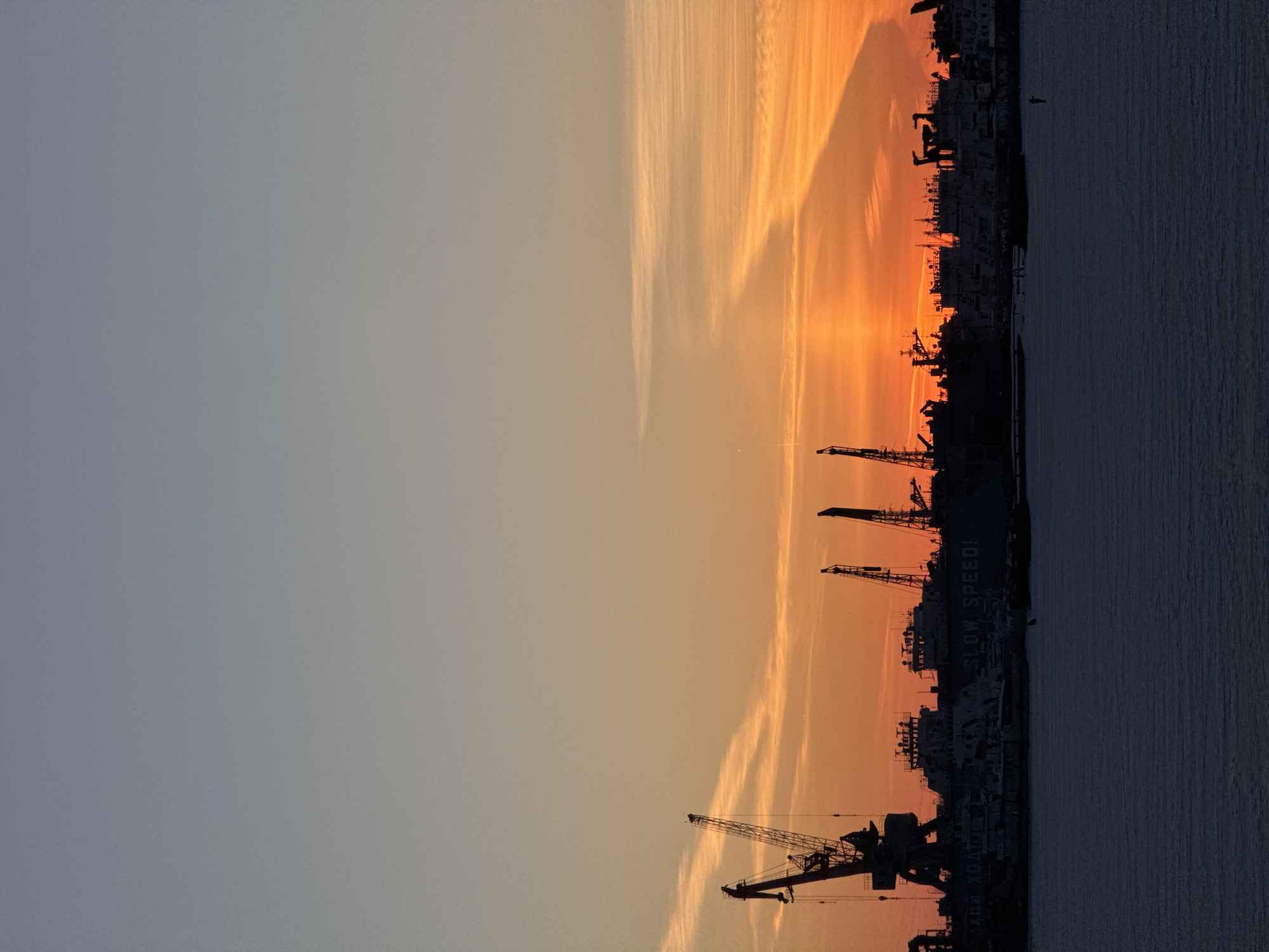 The image depicts an industrial waterfront scene in Chaikovsky, Russia, during either sunrise or sunset. The sky transitions from light grey-blue at the top to a vibrant orange-red at the horizon, featuring horizontal cloud formations and a distinct diagonal shadow cast across the orange segment. The midground presents a dark silhouette of a shipyard or port facility, populated by multiple large industrial vessels, some appearing to be cargo ships, with faint legible text such as "AGE LOADED" on a ship on the left and "SLOW SPEED!" on a ship in the center. Numerous cranes, including a prominent gantry crane on the far left, are silhouetted against the bright sky. The foreground consists of a dark, flat expanse of frozen water, likely a river or reservoir, exhibiting subtle textural patterns. A small, dark, vertical silhouette is visible on the frozen surface slightly to the right of the image center. No explicit human figures or actions are clearly discernible.