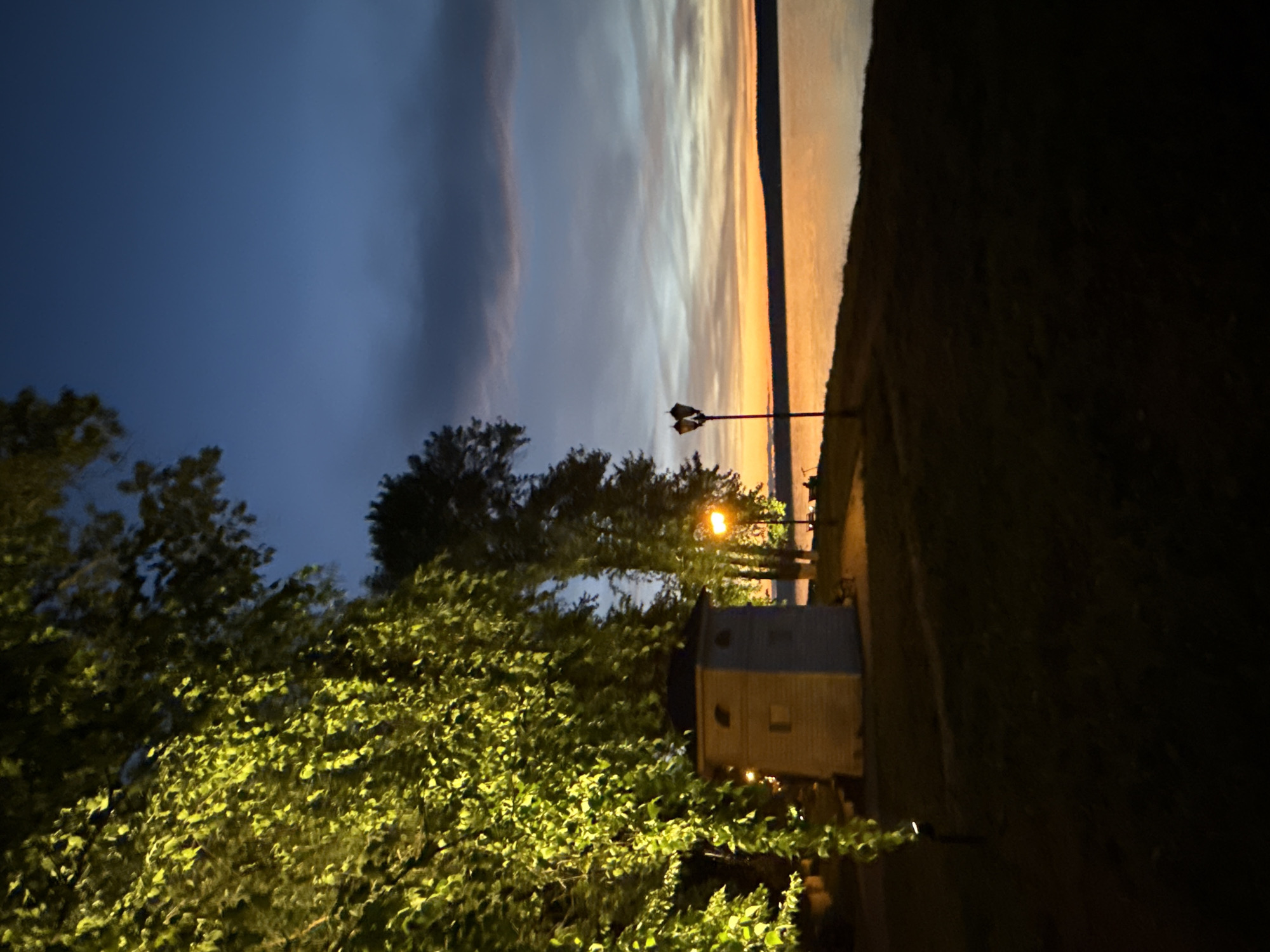 This image captures a twilight scene by a body of water, likely a reservoir or lake, in Votkinsk, Russia. On the left, the dense foliage of a tree, illuminated by artificial light, hangs over a small, light-colored building with a dark roof. The building features two arched windows and a rectangular window. Nearby, a lamppost with two globes stands.
The water reflects the warm orange and yellow hues of the setting sun, with a darker horizontal landmass visible in the distance. The sky transitions from a deep blue at the top to lighter shades near the horizon, with some clouds present. The foreground is a dark grassy area, suggesting a park or waterfront promenade. The overall atmosphere is tranquil, with the artificial lights adding a warm glow to the natural twilight setting. No people are clearly identifiable in the image, though a faint silhouette might be present on a bench near the water's edge on the right.