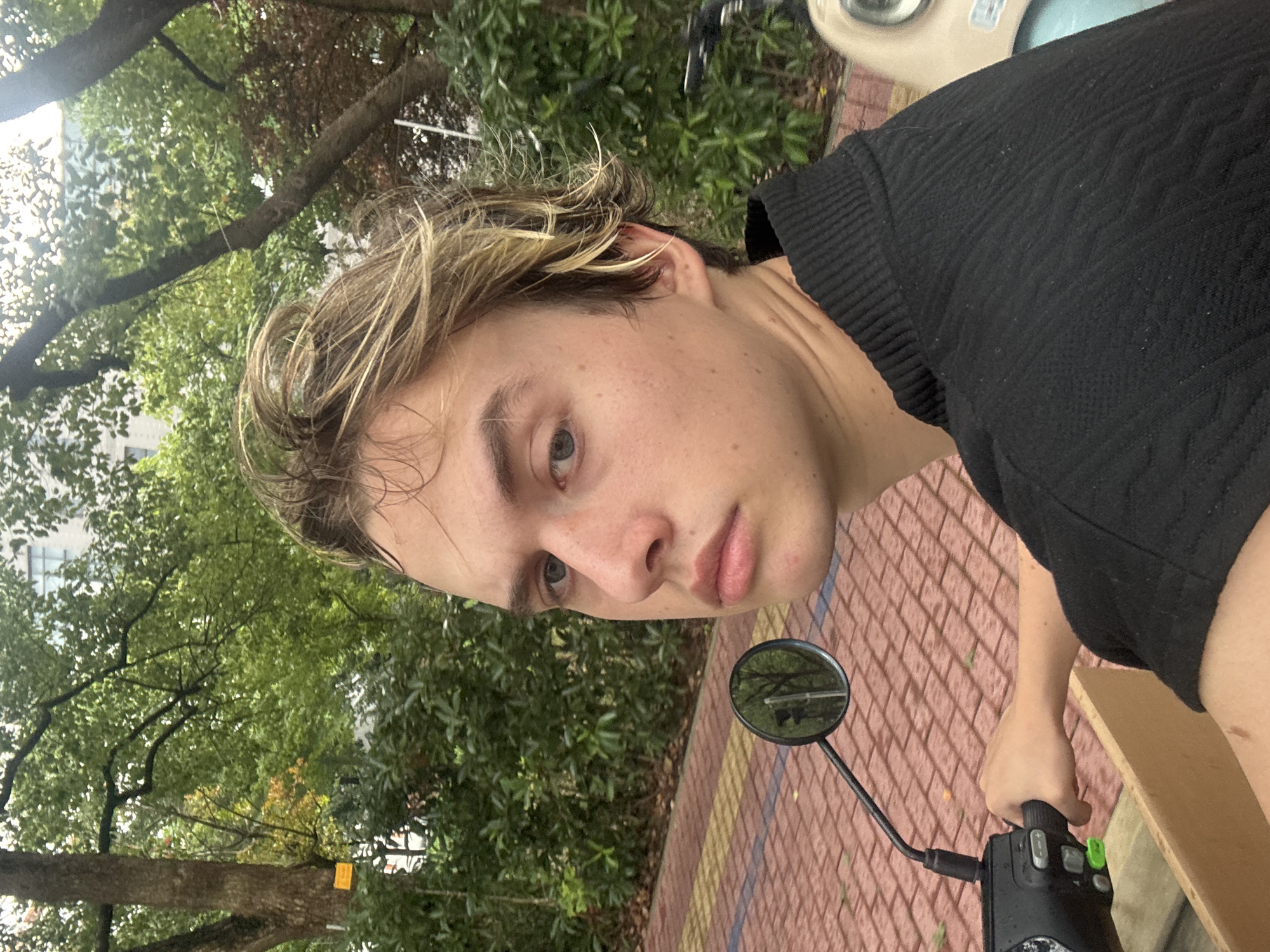 The image shows a young man with blond, messy hair sitting on a scooter in an outdoor setting, possibly a park or pathway in Suzhou Industrial Park, China. He is wearing a black, textured sweater and looking towards the camera with a neutral expression. His left hand is on the scooter's handlebar, which has green buttons. A circular mirror attached to the scooter reflects trees and a building. The ground is paved with reddish-brown bricks arranged in a pattern. Lush green foliage and trees are visible in the background, with a hint of a building in the distance. The overall lighting suggests an overcast day.