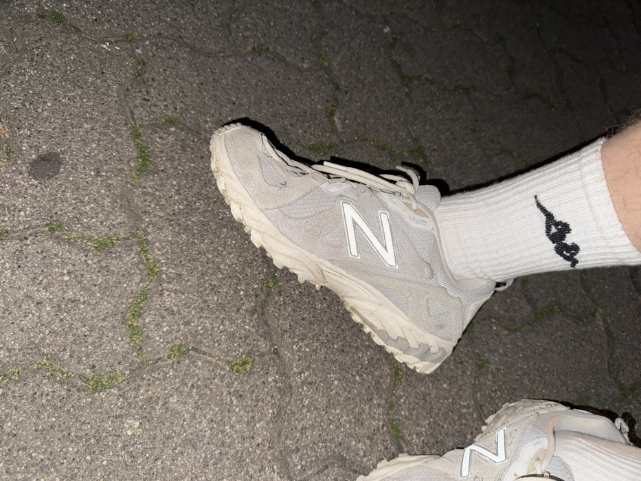The image displays a close-up, low-angle view of a person's lower legs and feet, resting on a dark grey, coarse-grained asphalt or concrete surface. The surface exhibits numerous cracks, within which green moss or small plants are growing in irregular patterns. The environment appears dimly lit, indicative of dusk or nighttime conditions.
The person is wearing white, crew-cut socks. The visible right sock features a black embroidered logo depicting a stylized quadrupedal animal, consistent with a dinosaur or reptile in a dynamic pose. Both feet are clad in light-colored, likely off-white or light grey, New Balance brand athletic shoes, identifiable by the distinct "N" logo on the lateral side of the right shoe. The footwear shows signs of moderate wear and minor soiling. The visible skin on the lower leg is light-toned and has visible hair. The image contains no intrinsic visual elements to confirm the specified location as Strasbourg, France.