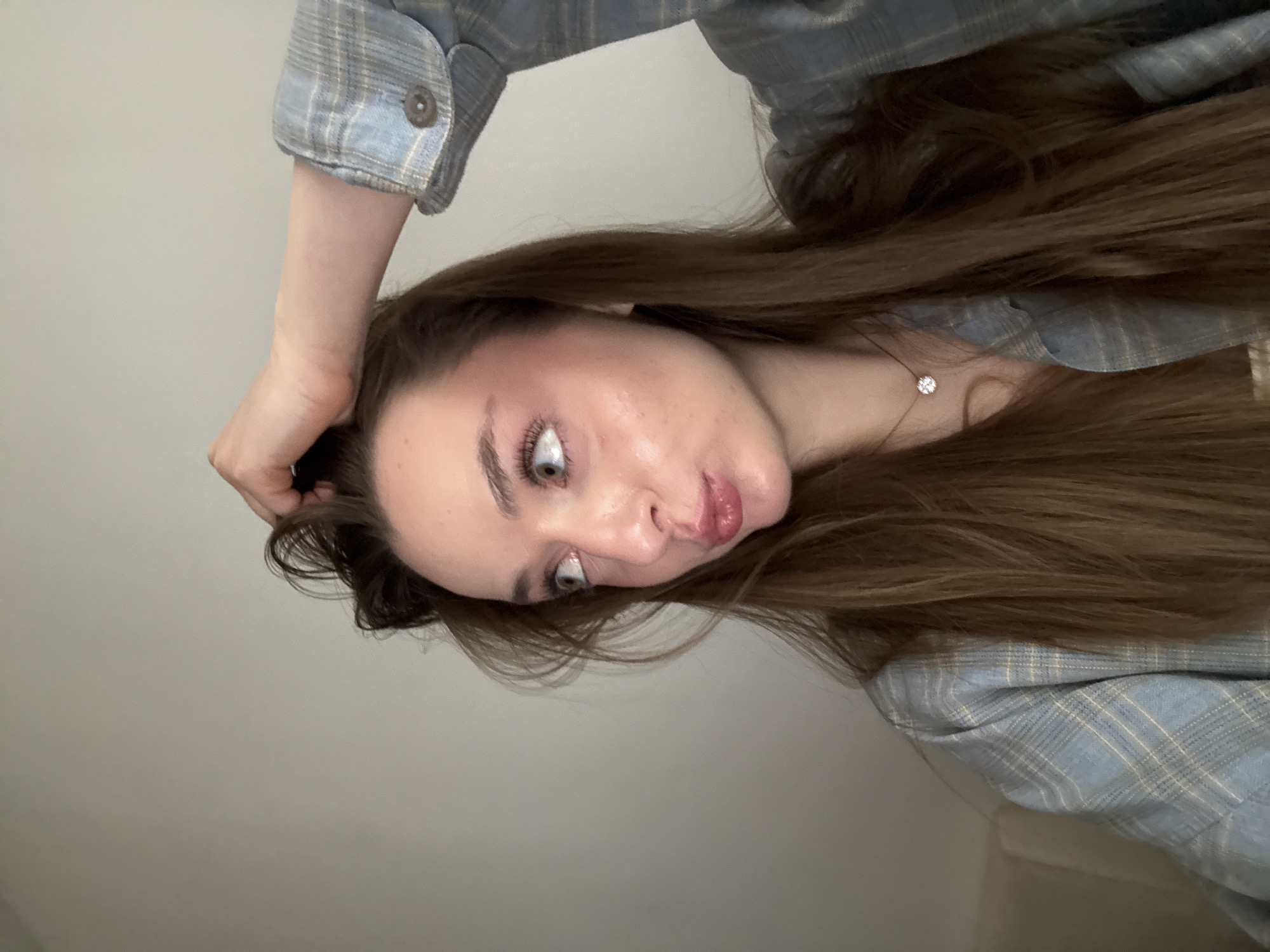 A woman with long brown hair is featured in a close-up shot. She is wearing a light blue plaid shirt and a delicate necklace with a small diamond. Her head is tilted to the side, with one hand resting on her hair. The background is a plain, light-colored wall. The image provides no information about the location in Kragujevac, Serbia, or any other details about a specific city or environment.
