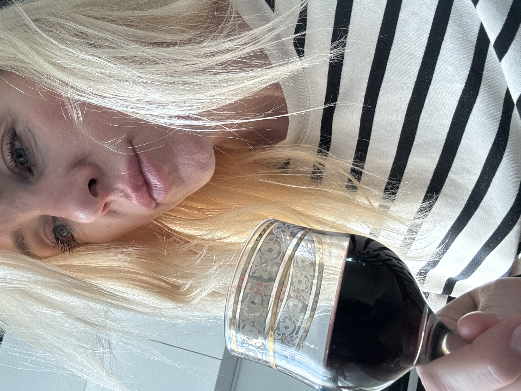 A close-up, high-angle shot shows a woman with blonde hair holding a glass of red wine. She is wearing a black and white striped shirt. Her face is partially obscured by her hair. The woman appears to be indoors, with a white wall visible in the background. The lighting is bright and appears to be natural. There are no other people or objects of note in the image. The location is Unknown, Russia.