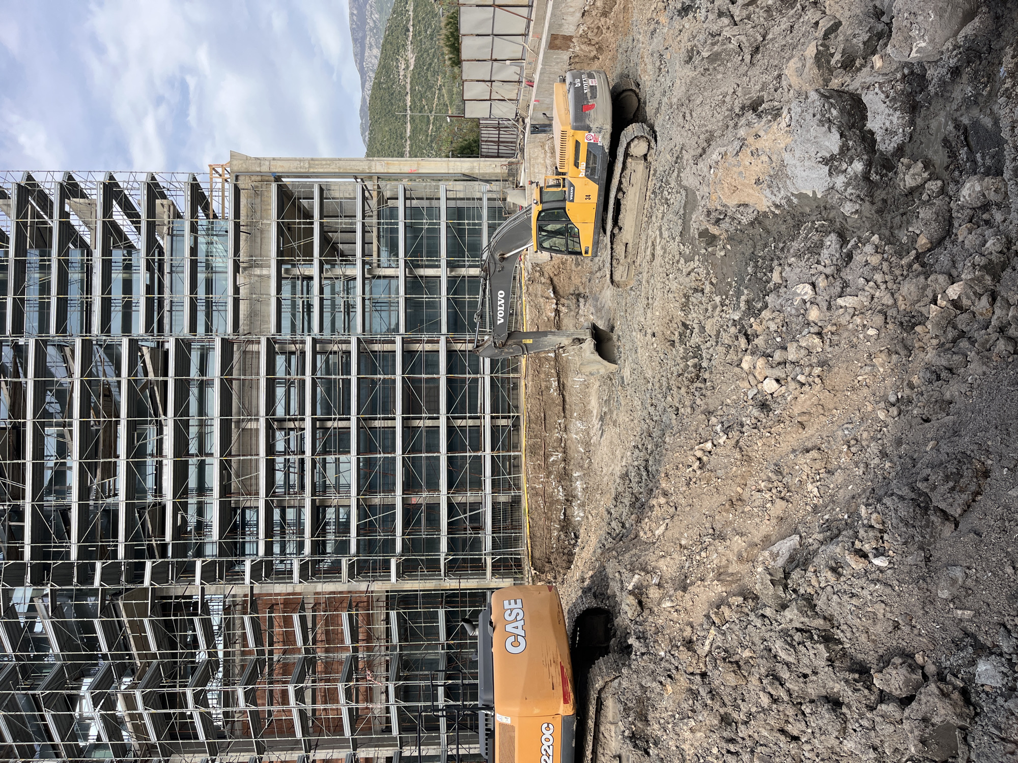 The image shows a construction site next to a building in Budva, Montenegro. A Volvo excavator is digging into the ground, while a CASE excavator is parked nearby. The building is under construction, with scaffolding covering its facade. A mountain is visible in the background.