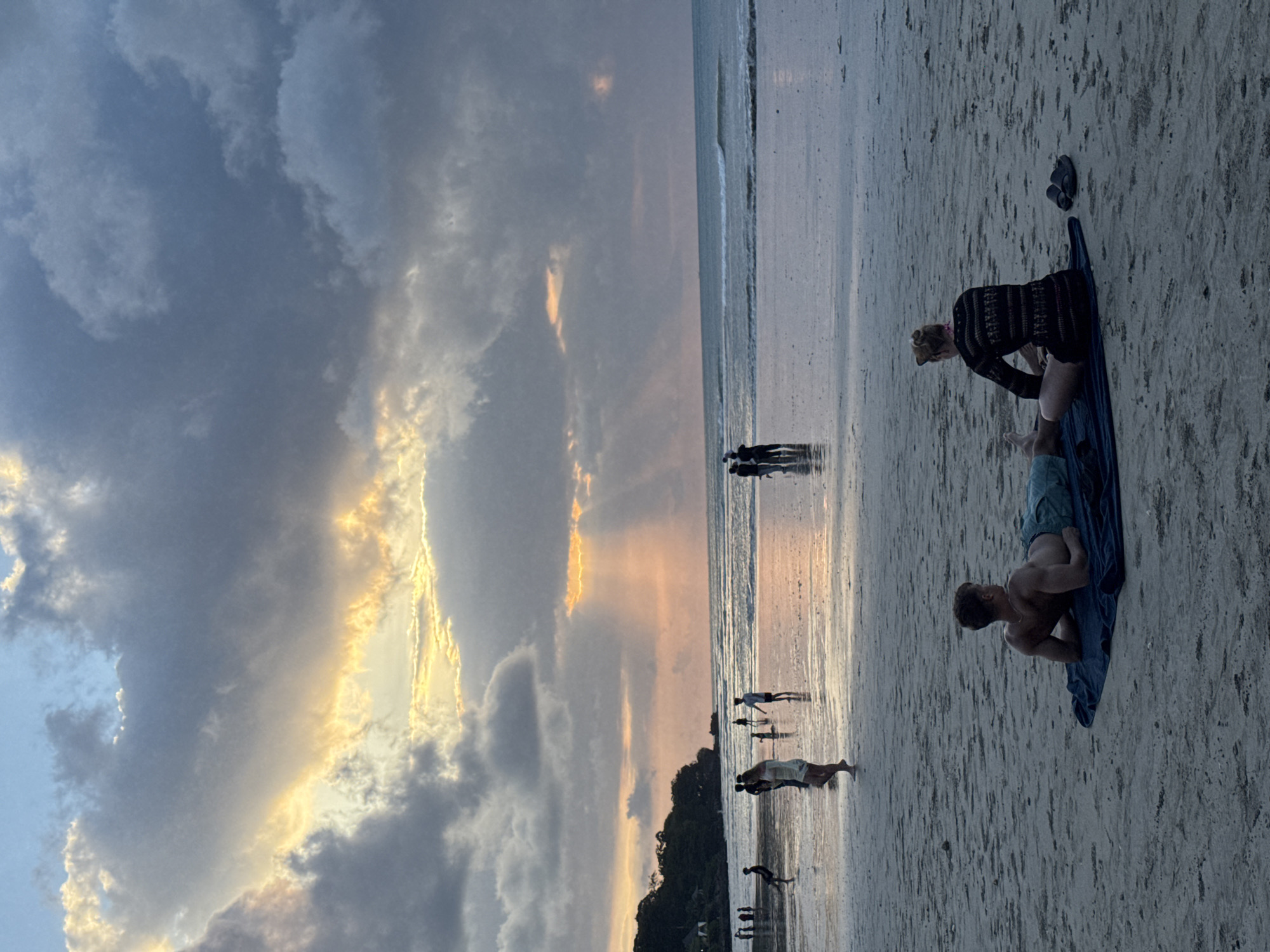 The image captures a serene beach scene during sunset or dusk in Kuta Selatan, Indonesia. The sky is filled with dramatic clouds, displaying a gradient from warm golden-orange hues near the horizon, illuminated by the setting sun, to cooler blues and greys higher up. The calm sea gently laps against a wide, reflective sandy shore, mirroring the sky and the silhouettes of people.
In the foreground, on the right, a man lies shirtless on his stomach on a blue towel, facing away from the camera and looking towards the water. Next to him, a woman with light hair, tied up, kneels or bends over on a similar blue mat. She is wearing a dark, possibly patterned, long-sleeved top. A pair of black flip-flops rests on the sand near her.
Further along the shoreline and in the shallow water, several small groups of people are visible, enjoying the evening. Some are walking along the wet sand, others are wading in the water, and one figure appears to be jumping or playing. Their forms are largely silhouetted against the bright reflections. A distant tree line or small hill is discernible on the left side of the horizon. The scene conveys a sense of peaceful relaxation, with individuals enjoying the beauty of the sunset by the ocean. No visible text is present in the image.