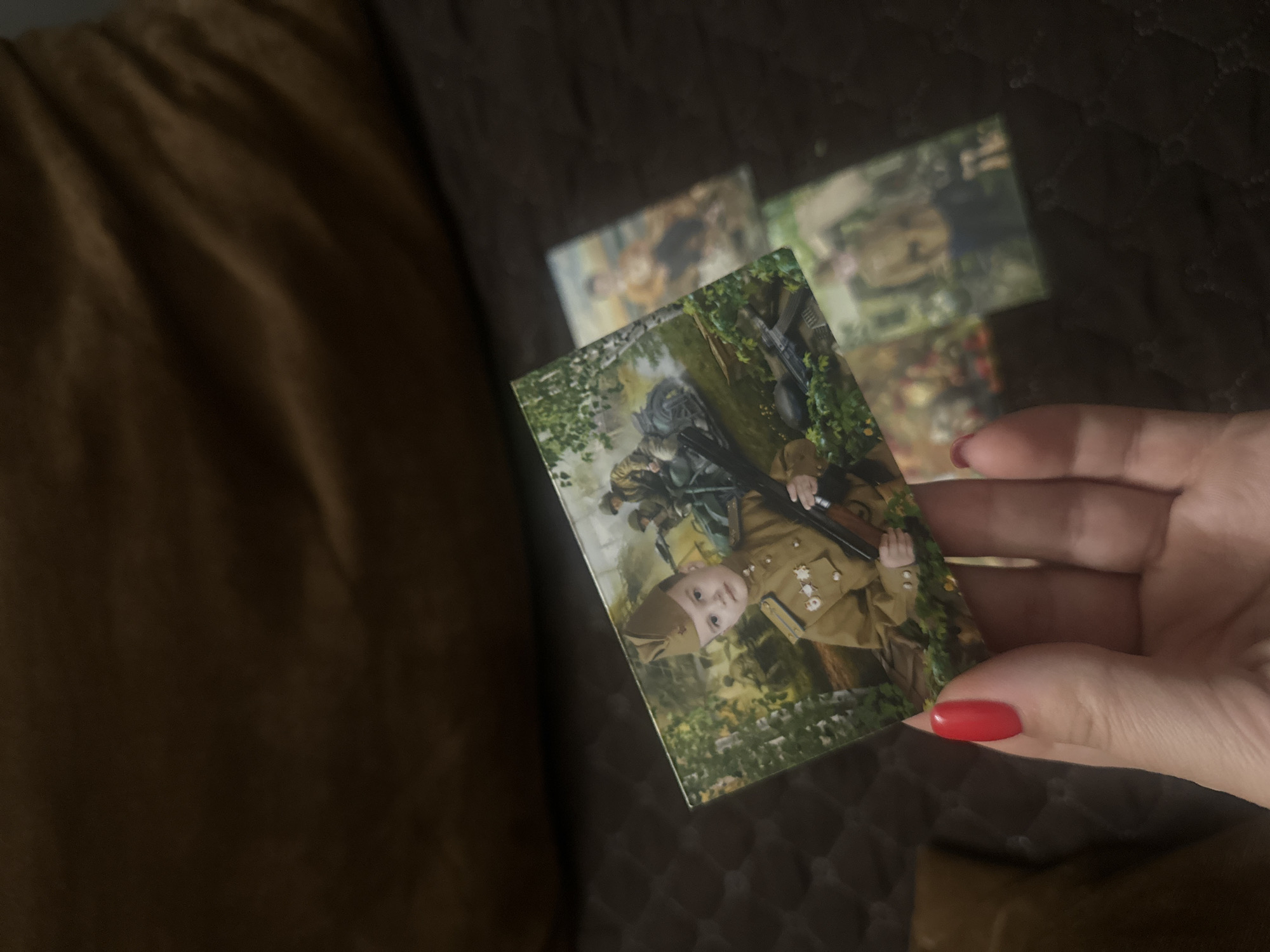 A hand with bright red nail polish is holding a photograph. The photograph depicts a young boy dressed in a military uniform, complete with a hat and medals, holding a rifle. In the background of the photograph, other soldiers and military vehicles are visible, suggesting a historical or reenactment scene. The overall tone of the photograph appears to be serious and commemorative. The hand and photograph are placed on a dark, textured surface, possibly a couch or blanket.