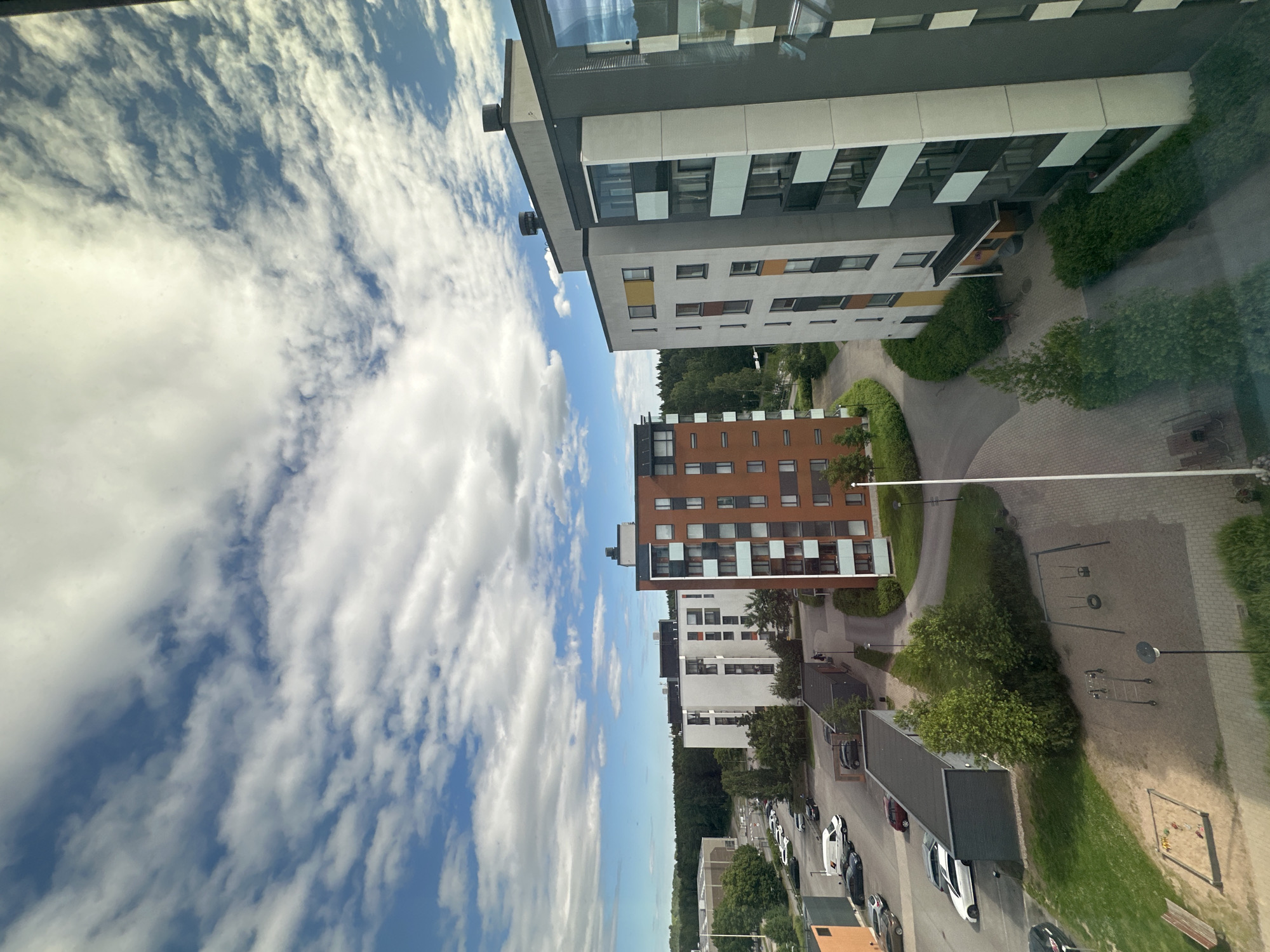 An aerial, eye-level shot captures a residential neighborhood with several apartment buildings and a playground under a partly cloudy sky. In the foreground on the right, a tall, modern apartment building with a dark grey facade and white vertical accents dominates the frame. To its left, a shorter, reddish-brown brick apartment building with white-framed windows is visible. Further back, a row of white apartment buildings with black trim and balconies are arranged along a curved road. To the far left, a large, light-colored industrial or commercial building stands adjacent to a dense forest. A playground with swings, a sandpit, and climbing equipment is situated in the lower-left quadrant of the image, adjacent to a parking lot filled with several cars and a white van. The ground is a mixture of paved pathways and green grass.
