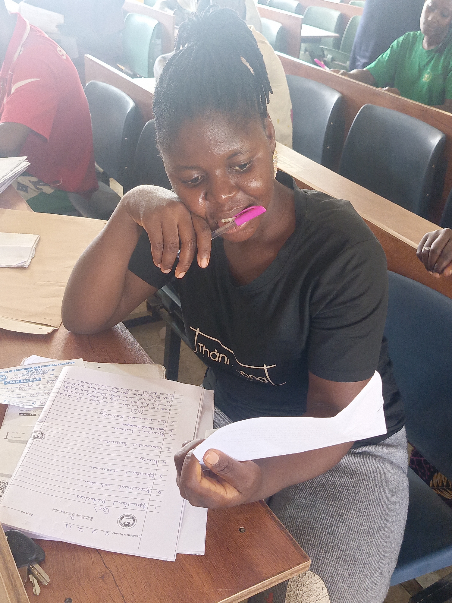 A young Black woman with dreadlocks, wearing a black t-shirt and grey skirt, is seated at a wooden desk in what appears to be a classroom or lecture hall in Akwanga, Nigeria. She is the central focus, looking down intently at a notebook and loose papers, with a pink pen held in her mouth, suggesting deep thought or concentration. Her left hand rests on the table, while her right hand holds a folded white paper.
In the background, other individuals are partially visible, seated at similar desks and chairs; one person wears a red shirt to the left, and another in a green shirt is visible further back to the right. The setting features rows of brown wooden desks and dark-colored chairs.
The main subject is engaged in an academic activity. Her notebook contains handwritten notes related to various agricultural subjects, including "Agricultural production," "Agricultural extension," "Forestry," "Ornamental horticulture," and "Food Science and technology." The notes also define "Ornamental Horticulture" and list examples of crops. A "Candidate's Number 22211" is visible at the top of the page. A "CASH RECEIPT" from the "SCHOOL OF VOCATIONAL AND TECHNICAL EDUCATION," "COLLEGE OF EDUCATION, AKWANGA" is also visible on the desk, indicating the educational context and location. The receipt shows an amount of "N127.50." Keys are also on the desk to the left. The lighting suggests it is daytime.