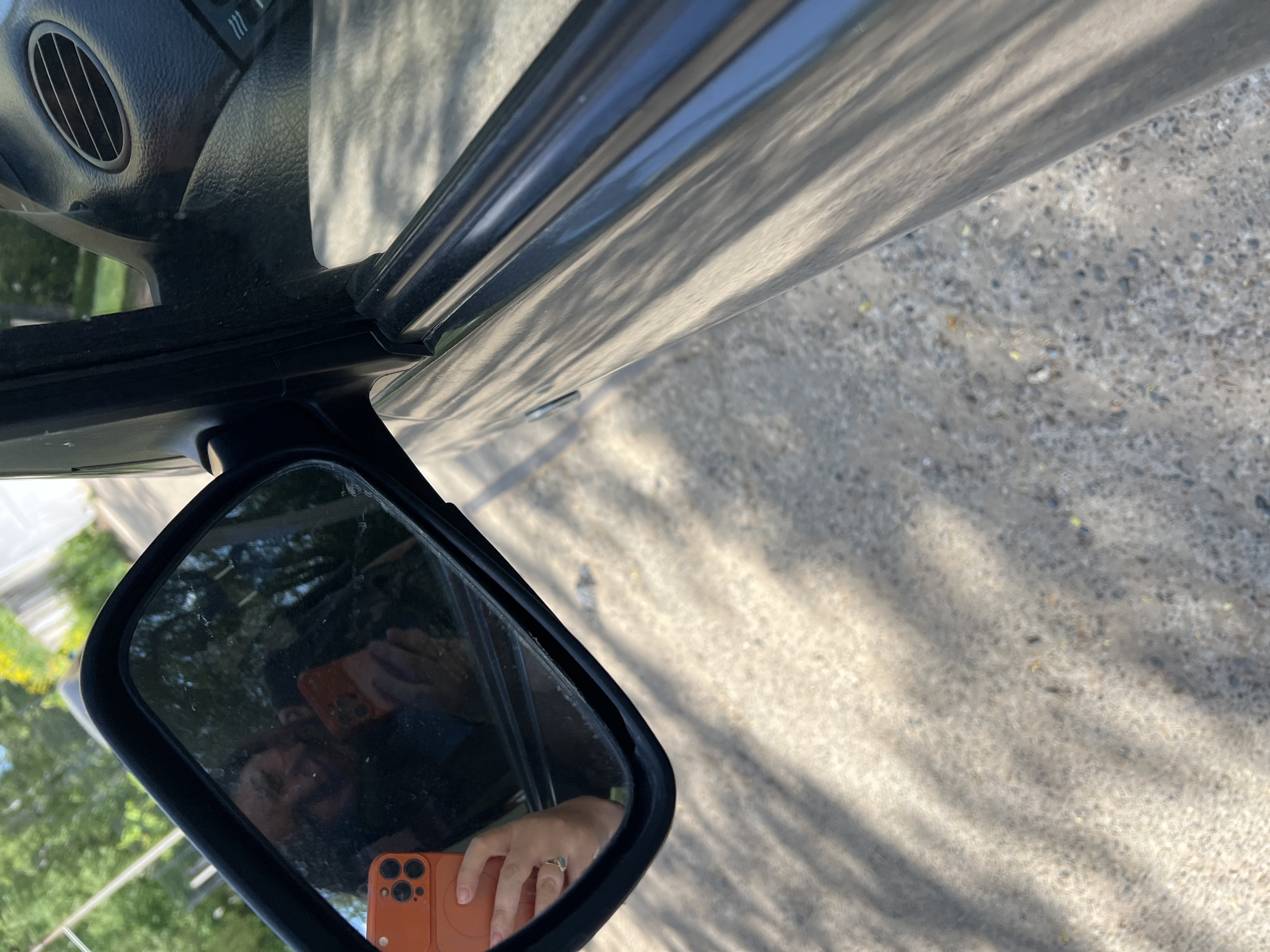 The image presents a close-up, obstructed view from inside a vehicle, possibly a car, with a side mirror being the primary focus. The reflection in the side mirror shows a person holding an orange smartphone, presumably capturing the image. The background reflected in the mirror indicates a setting with trees and vegetation, suggesting an outdoor environment. Outside the car, the ground appears to be paved, potentially a street or parking area. No clear indicators definitively locate the scene in Noviy, Russia.