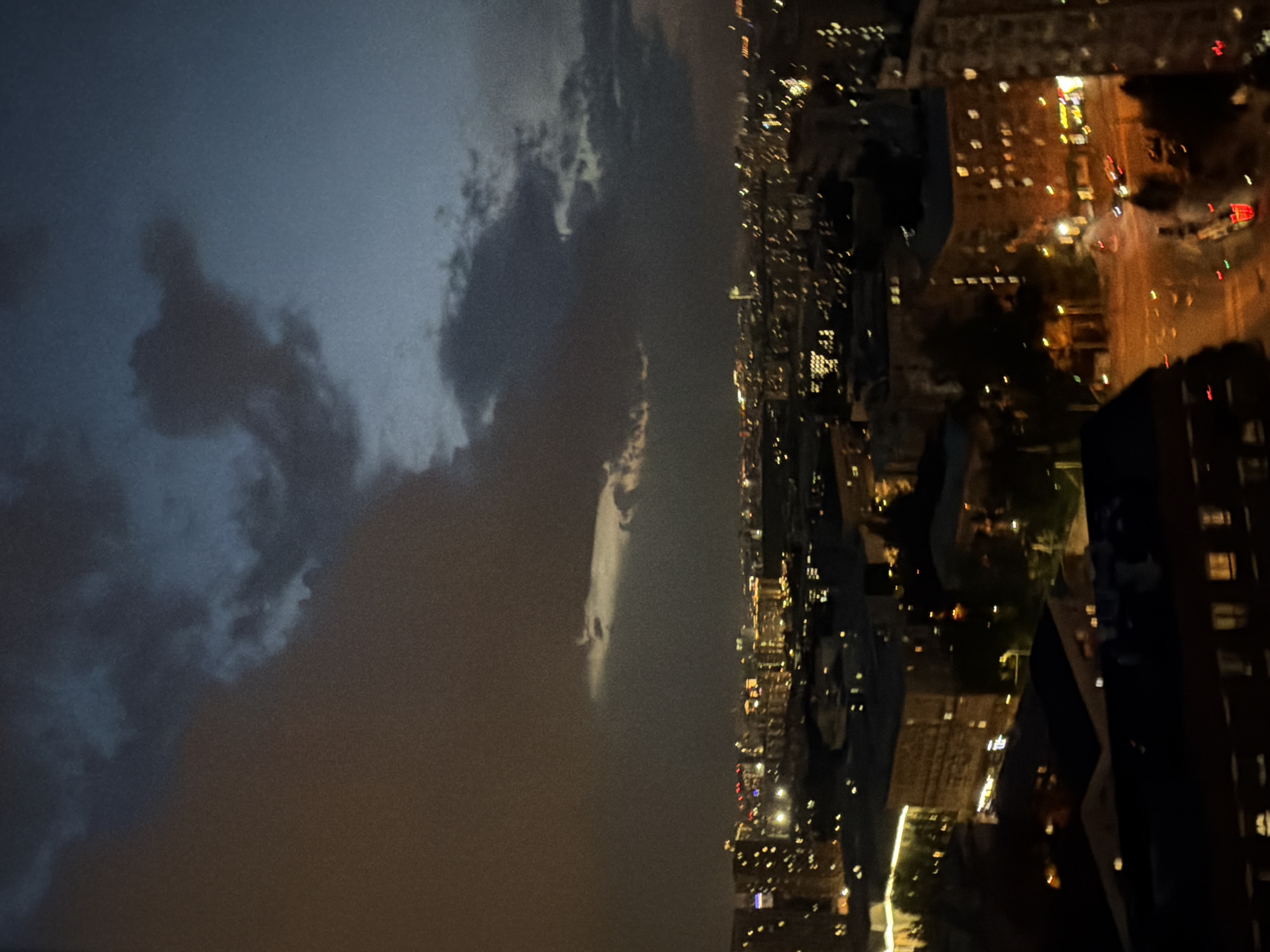 The image shows a nighttime view of an urban area, likely taken from a high vantage point. The sky is filled with dark clouds, some illuminated from below. The cityscape is visible with numerous buildings lit up, and the blurred lights of traffic can be seen on the streets. The overall environment suggests a city at night during or just after a storm.