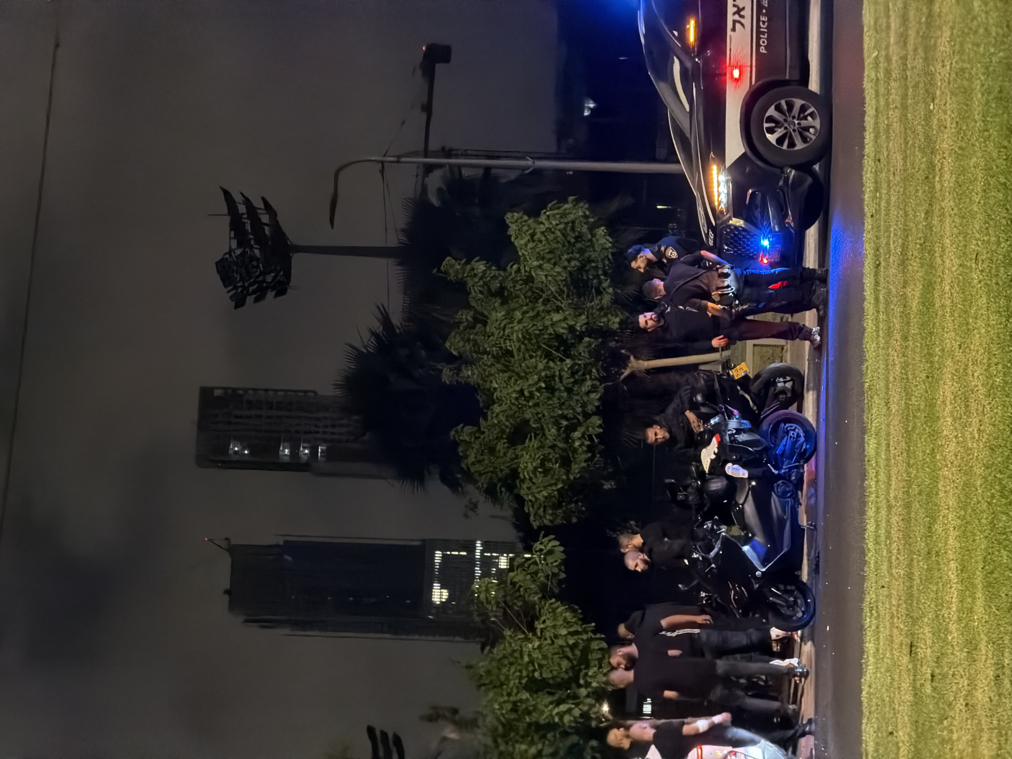 At night in Tel Aviv, Israel, near a busy street with a grassy median, several people are gathered. Two police officers in uniform, one with a vest and flashlight, stand near a police SUV with flashing blue and red lights. The SUV has the word "POLICE" visible on its side. Several men, some in dark clothing, are also present, standing near motorcycles. One motorcycle is black and parked in the foreground. In the background, tall buildings and palm trees are visible under a dark, hazy sky. The overall scene suggests a nighttime incident or investigation.