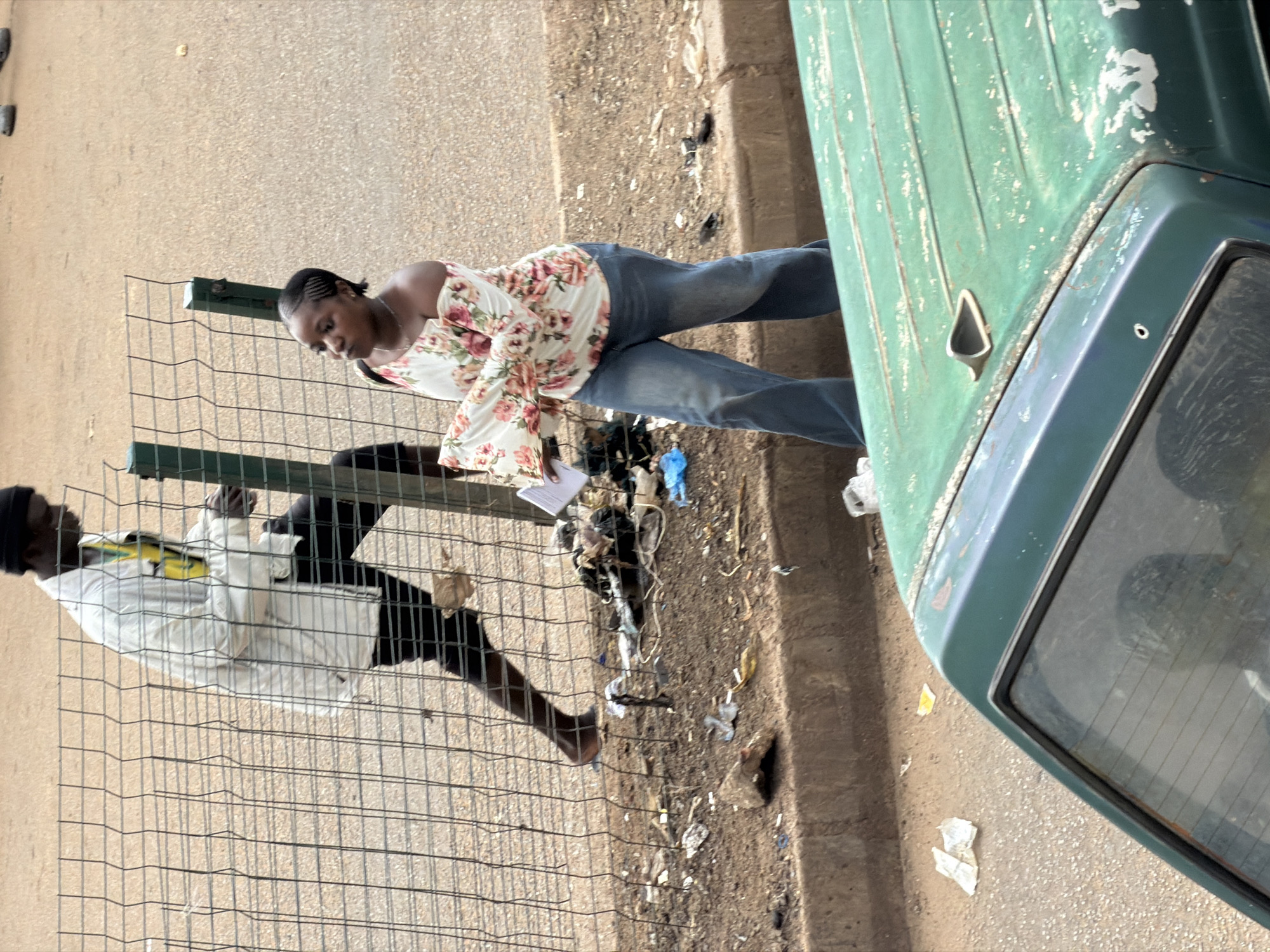 In Bukuru, Nigeria, a man wearing a white jacket and black pants walks near a metal fence. A woman with braided hair is also present, wearing an off-the-shoulder floral top and blue jeans. She is holding some papers. The ground is littered with trash, and a green vehicle is partially visible in the foreground.