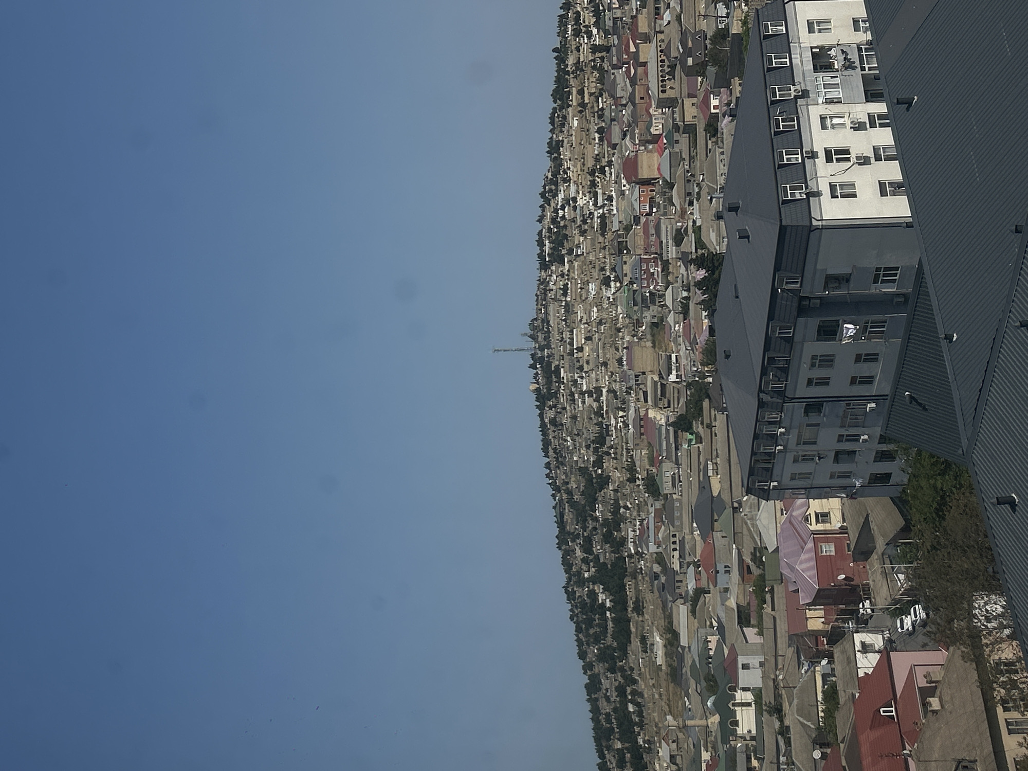 This is a high-angle, medium shot of a city on a hillside under a clear blue sky. The city, identified as Khyrdalan, Azerbaijan, is densely populated with a mix of residential buildings. In the foreground, a dark gray, modern apartment building with multiple windows and air conditioning units is prominent. Behind it, a variety of houses with different roof colors, including red, pink, and green, are visible. The hillside in the background is covered with more buildings and dotted with trees. A television tower and what appears to be a religious structure are situated on the highest point of the hill. The scene depicts a residential urban landscape.