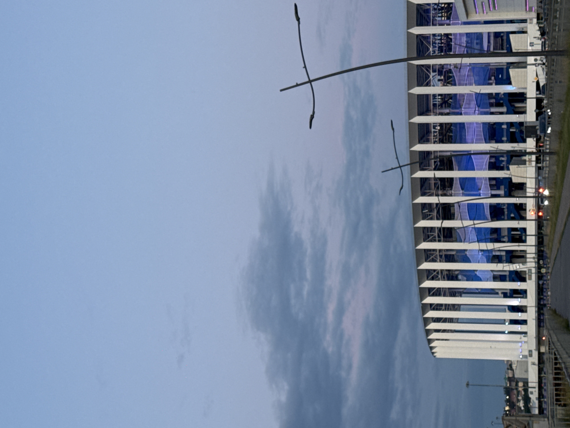 This is a vertically oriented, medium-distance, eye-level shot of a modern, white, multi-story building with blue accents, seen from the side. The building's facade is characterized by horizontal white louvers that extend outwards. Behind the louvers, translucent blue netting or fabric is visible, suggesting it might be a covering or part of the building's design. The building appears to be an arena or stadium, with visible structural elements and lighting.
In the foreground, to the left of the building, is a clear, pale blue sky with some wispy, darker clouds. Two tall, thin lampposts with multiple light fixtures are prominent in the sky. One lamppost is positioned in the upper left quadrant, and another is closer to the building on the right side of the image. The overall impression is that of an outdoor setting, likely in the evening, given the twilight sky and the artificial lighting on the building and lampposts.
To the right of the building, a road or pathway is visible, with traffic lights and what appear to be vehicle lights, indicating activity. A sidewalk or pedestrian area runs alongside the road. In the far distance, beyond the building, there are glimpses of other structures and lights, suggesting an urban or developed area.
The image appears to have been taken in Nizhny Novgorod, Russia, based on the provided location context. The time of day is likely dusk or early evening, as the sky is not fully dark, and artificial lights are beginning to illuminate the scene. The weather appears clear with scattered clouds. No people are clearly discernible in the image. The scene conveys a sense of modern architecture and urban infrastructure. There is no visible text in the image.