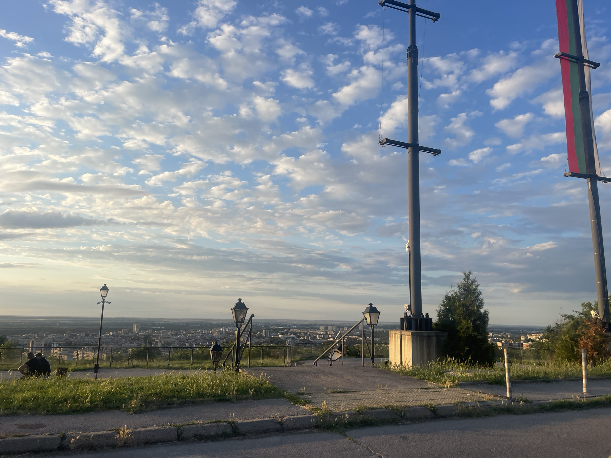 The image depicts a scenic view overlooking the city of Ruse, Bulgaria, from an elevated vantage point. In the foreground, a paved area with grass and a sidewalk leads towards a railing overlooking the cityscape. Several lampposts are visible, adding to the atmosphere. Two individuals are seated on the grass to the left, appearing to observe the view.
Dominating the right side of the frame are two tall, dark poles, one of which displays a flag with horizontal stripes of red, white, and green. These poles appear to be part of a significant structure or monument. A small evergreen tree stands between the two poles, closer to the right pole. The background reveals a sprawling urban landscape of Ruse under a sky filled with scattered clouds. The lighting suggests it is either early morning or late afternoon, with the sun casting a soft glow. The perspective from this vantage point highlights the city's spread and its relationship with the surrounding terrain.