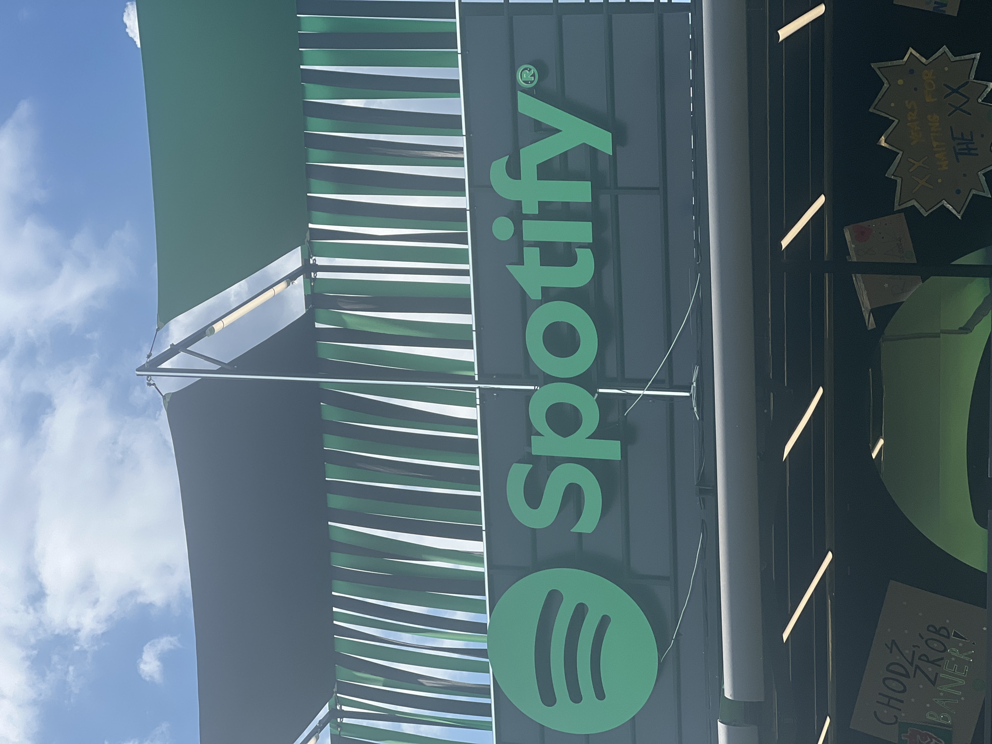 The image shows a close-up of a Spotify logo on a dark, vertical surface, likely a building facade. The logo, in its characteristic bright green color, is prominently displayed and includes the Spotify "swoosh" icon. To the left of the logo, there's a striped awning or facade element with alternating dark green and black stripes. Above, the sky is visible with some clouds, suggesting it's daytime.
In the background, a glimpse of other signs or posters can be seen. One sign, with a starburst shape, has some text on it. Another, to the lower right, appears to be a handwritten sign with Polish text. The overall scene is an outdoor setting, possibly a street or an open area. The lighting suggests it's daytime, and the presence of clouds indicates typical weather conditions. No people are visible in the frame. The image is oriented vertically.
Visible text includes:
- The Spotify logo
- Polish text on a sign in the lower right: "CHODŹ ZRÓB BANER!" (roughly translated to "Come on, make a banner!")
- Text on the starburst sign: "XX YEARS WAITING FOR THE XX"