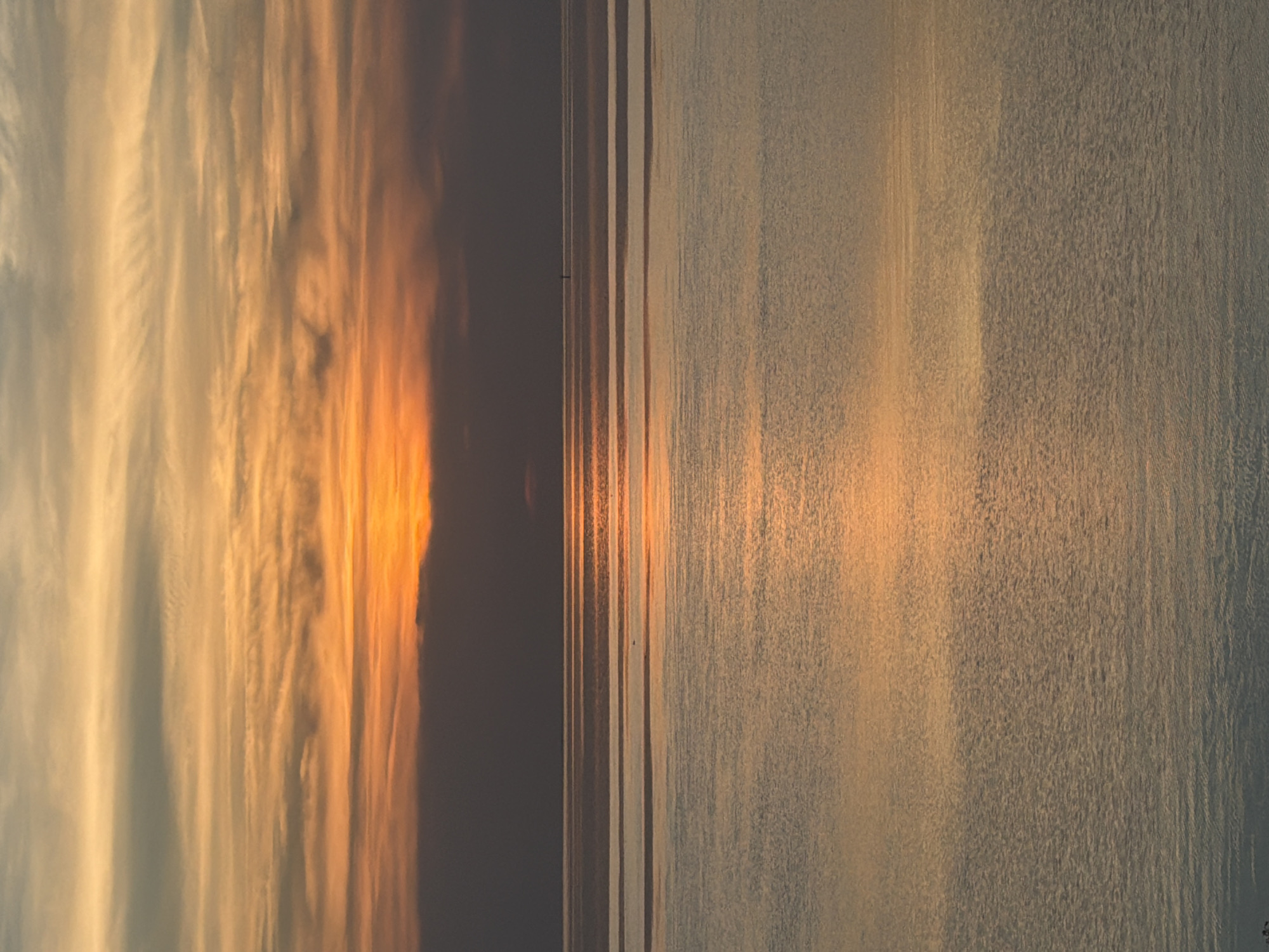 The image depicts a serene seascape at dusk. The sky is a canvas of warm colors, with layers of orange, yellow, and muted grey clouds stretched horizontally. The sun, a bright orange orb, is partially obscured by a dark band of clouds near the horizon. Below the sky, the calm sea reflects the celestial display, with horizontal bands of light and shadow creating a rippling effect. A thin, dark line of what appears to be a distant landmass or a very low-lying cloud is visible on the horizon. In the middle distance of the water, a small, dark, vertical marker or buoy stands out against the reflections. The overall atmosphere is tranquil and visually striking, characteristic of a sunset over a body of water.