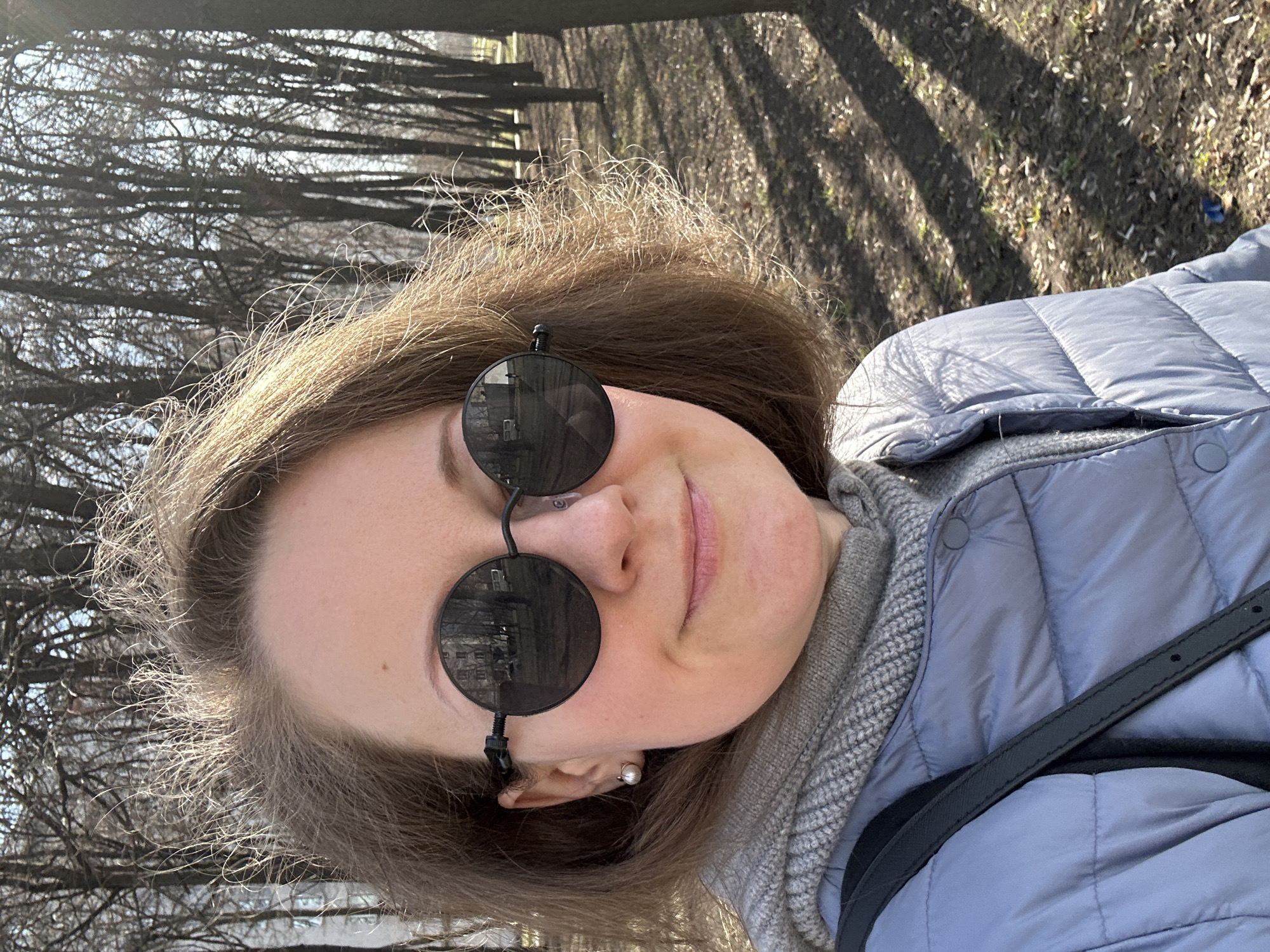 A woman is taking a selfie in what appears to be a park. She has fair skin, light brown hair, and is wearing round, dark sunglasses. She has a gray scarf and a blue quilted jacket on. A black strap is visible, crossing her chest. The background shows a park setting with bare trees, casting long shadows on the ground. Based on the architecture reflected in the sunglasses and the style of trees, the location is likely in a park in Moscow, Russia.