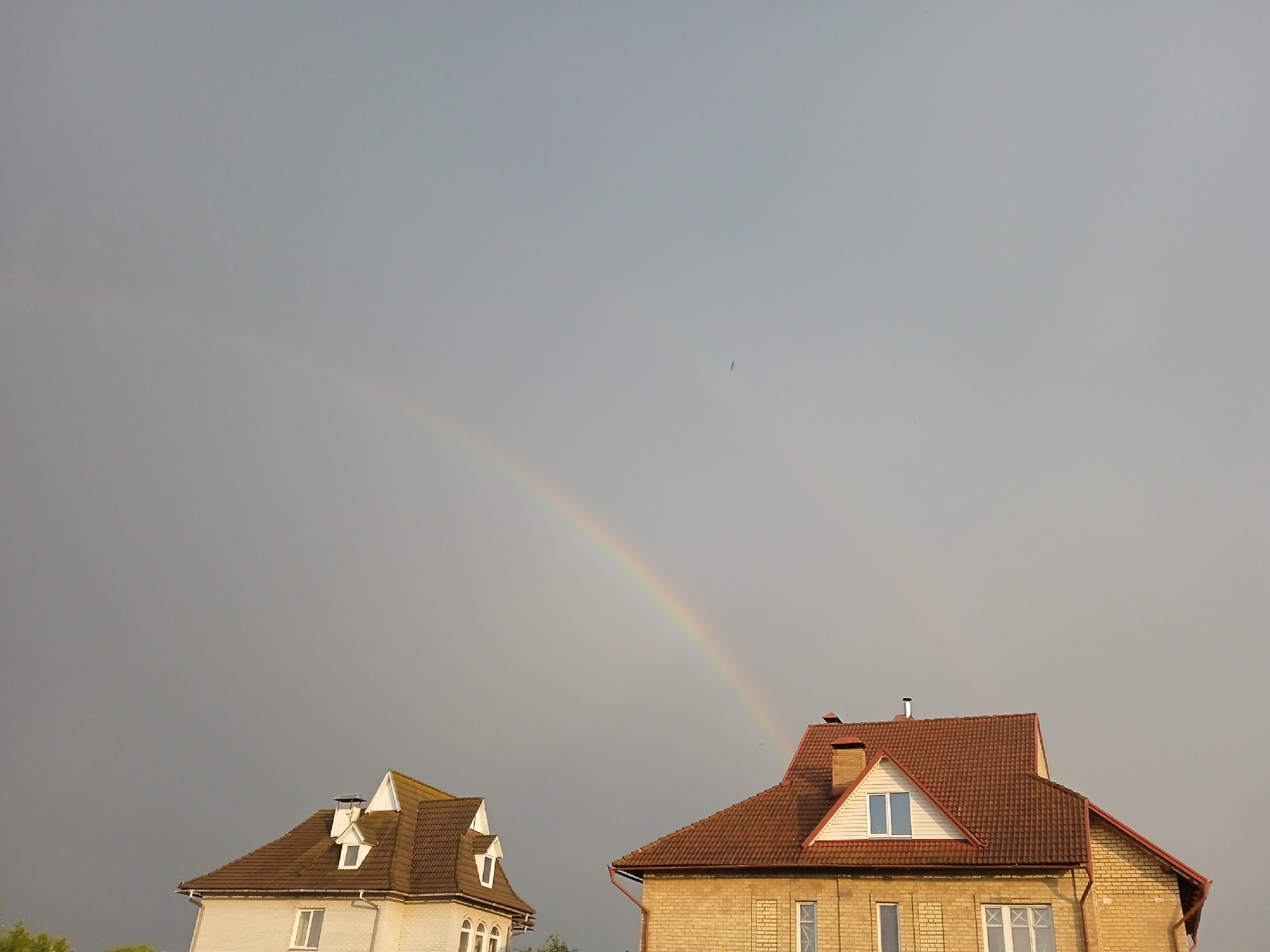 The image shows a dull, overcast sky with a faint double rainbow arching across it. In the foreground, two houses with reddish-brown tiled roofs are visible. The house on the left is lighter in color, possibly white or light beige, with dormer windows. The house on the right is made of yellow brick and also features dormer windows. A chimney is present on the roof of the yellow brick house. The scene appears to be a residential area, likely in the city of Vyshhorod, Ukraine, given the context. No people are visible in the image. The lighting suggests it might be late afternoon or early evening, with the overcast sky and rainbow indicating recent or ongoing rain.
