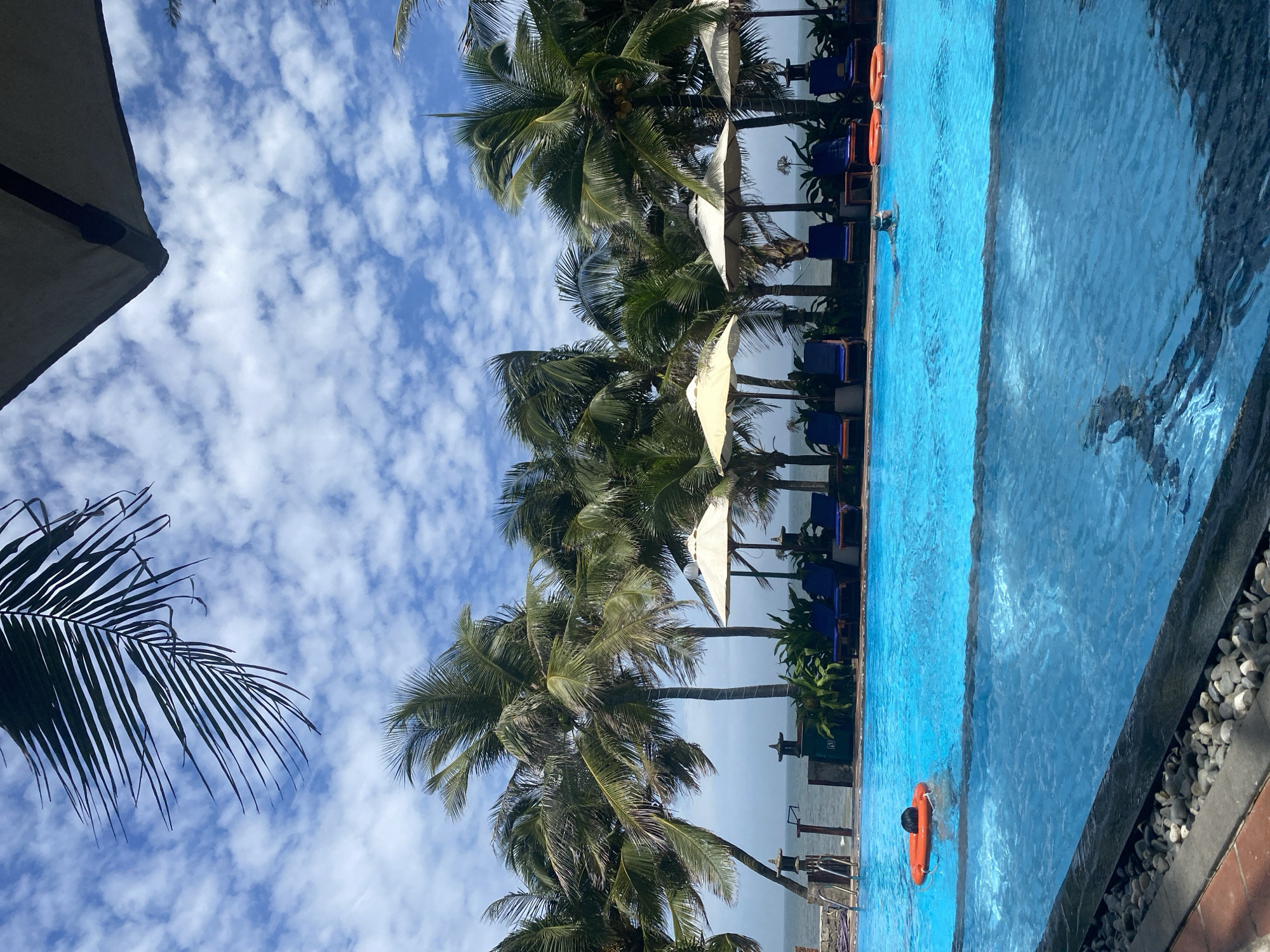 This is a vertical, eye-level, medium shot taken outdoors at a resort in Mũi Né, Vietnam. The foreground features the edge of a bright blue swimming pool with a dark gray coping stone and a border of pebbles and red tiles. In the pool, a person is swimming, wearing goggles and a swim cap, with their head and shoulders visible above the water. To the left, an orange life preserver floats near the surface. The midground is dominated by a row of lounge chairs and beach umbrellas, positioned behind the pool and in front of a line of palm trees. The palm trees are tall and lush, with their fronds swaying gently. Beyond the trees, the ocean is visible, with a slightly hazy horizon. The sky above is a vibrant blue, filled with scattered white clouds. The overall scene depicts a tropical resort setting on a bright, sunny day.