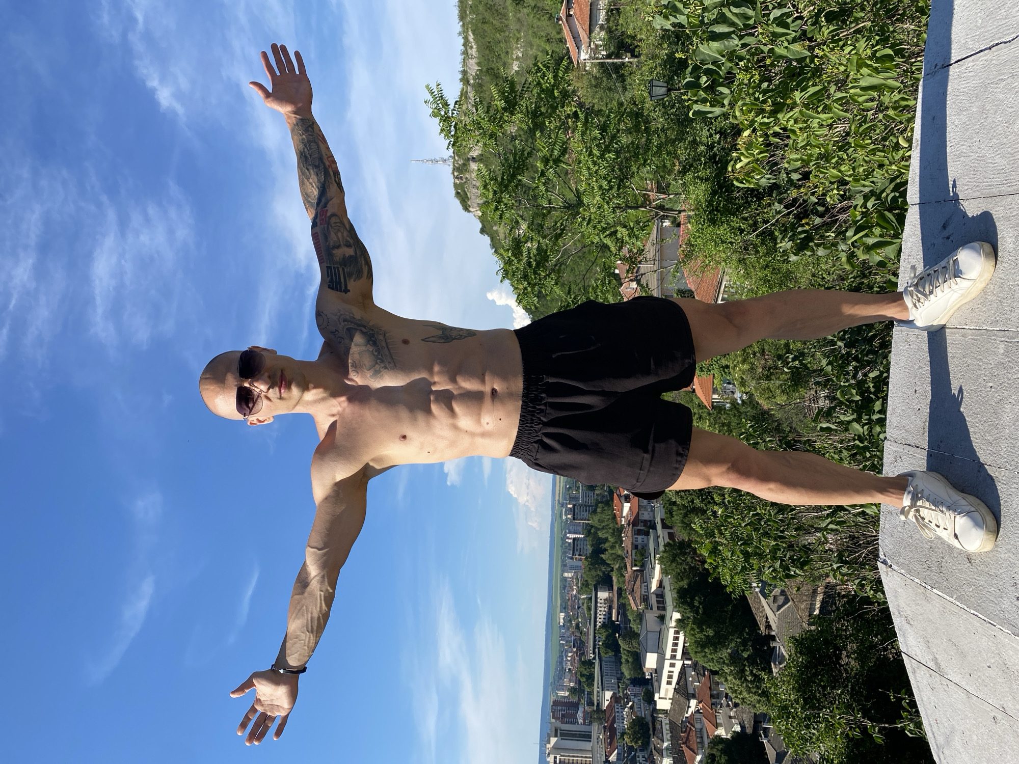A shirtless, bald man wearing sunglasses and black shorts stands with his arms outstretched, facing the camera. He has a muscular physique and visible tattoos on his arms. He is standing on a light-colored, flat surface, likely a stone or concrete platform, with a city and a hilly landscape in the background under a blue sky with some clouds. The city is identifiable as Lovech, Bulgaria, and a prominent feature in the background is the Lovech fortress and the TV tower located on Hisarya Hill. Lush green vegetation is visible around the platform and further up the hill.