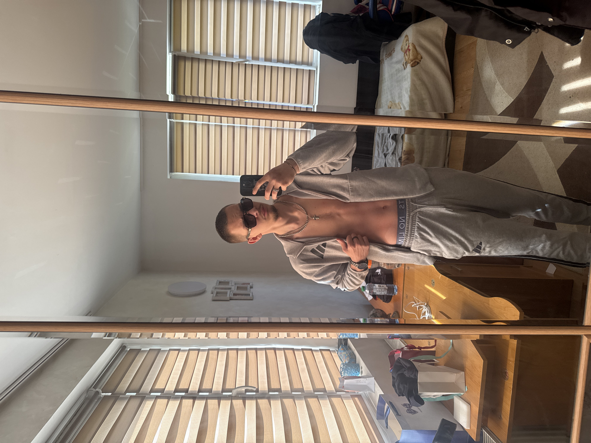 A man stands in front of a mirror taking a selfie. The man is wearing sunglasses, a gray tracksuit jacket that is open revealing his bare chest and silver necklace, and tracksuit bottoms. He is holding a black phone in his left hand to take the picture. The background features a window with horizontal blinds, a desk with items on it, and a bed with clothes and a patterned rug. The location is Vetren Dol, Bulgaria, based on metadata.