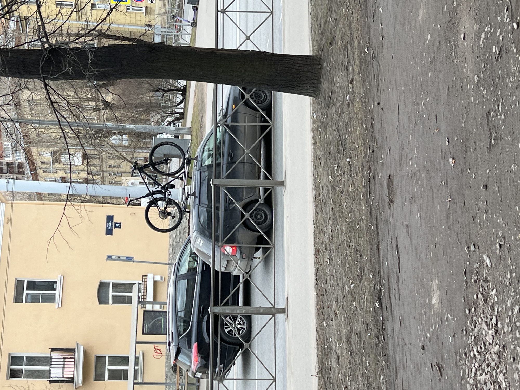 The image shows a street scene in Saint Petersburg, Russia, identifiable by the street sign "Ulitsa Sadovaya 34" on the building. A black bicycle is locked to a metal fence near a tree. Behind the fence, two black cars are parked close to each other. The street is visible to the right of the fence. The building is painted yellow and has multiple windows and a balcony. The tree is bare, suggesting it is either late autumn or winter.