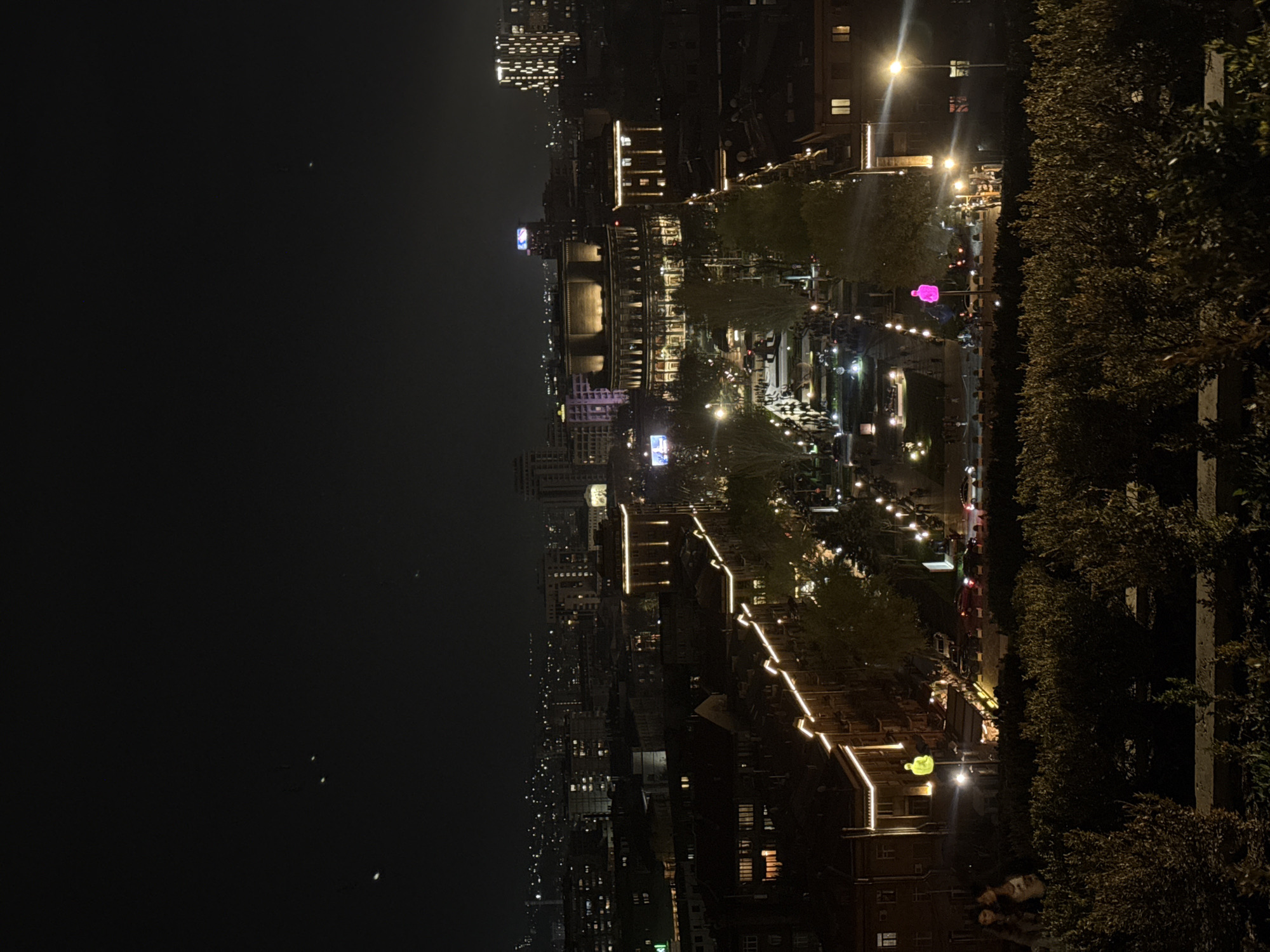 Captured at night, this high-angle, long-distance shot showcases an illuminated street scene in Yerevan, Armenia. The street, lined with trees and bordered by buildings, is brightly lit with streetlights and building lights. The buildings exhibit architectural details and are illuminated with warm lighting. People are visible walking along the street, and cars are moving through the scene, with their headlights and taillights creating light trails. The environment is urban, with a mix of residential and commercial buildings. The presence of lush trees adds an element of nature to the cityscape. In the background, more buildings and lights stretch into the distance. The overall atmosphere is vibrant and bustling, capturing the essence of Yerevan at night.