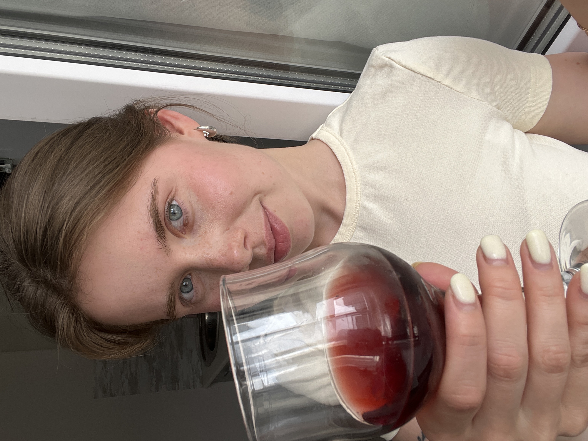A young woman is holding a glass of red wine. The setting is indoors, near a window. The location cannot be determined based on the image.