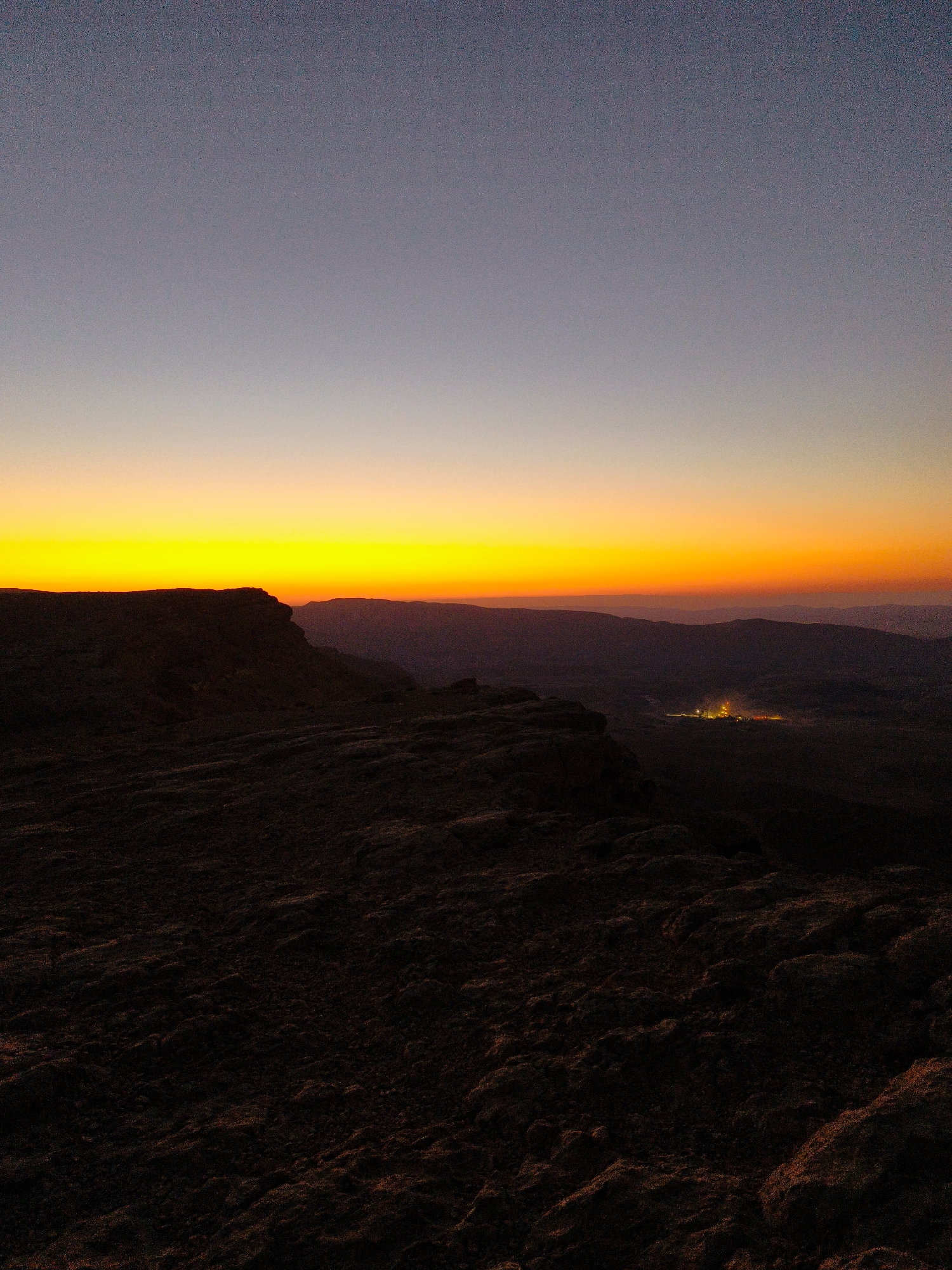 This image depicts a sunset or sunrise over the desert landscape surrounding Yeruham, Israel. The sky is a gradient of colors, transitioning from a deep blue at the top to a vibrant yellow and orange near the horizon. In the foreground, dark, rocky terrain suggests a high vantage point. In the middle ground, the silhouette of a cliff or plateau edge is visible. Further in the distance, a cluster of lights, likely representing the city of Yeruham, can be seen nestled among rolling hills, glowing against the darkening landscape. The overall scene conveys a sense of vastness and natural beauty.
