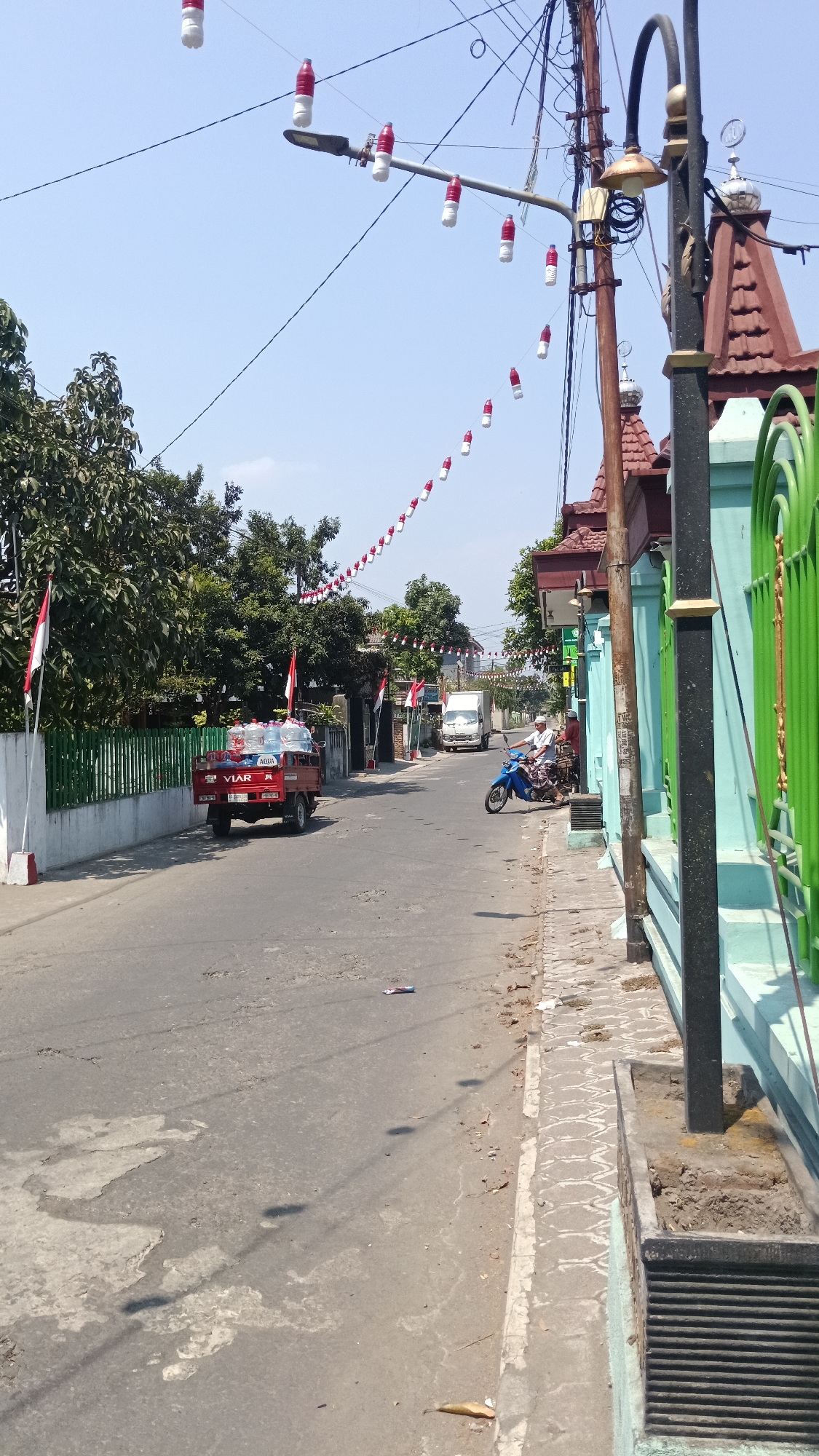 The image depicts a street scene in Purwokerto, Indonesia, likely on a clear, sunny day. The road is paved with asphalt and shows signs of wear, including patches and cracks. Along the right side of the road, there is a sidewalk with decorative patterned tiling, bordered by a light blue wall and a green metal fence. A dark lamppost stands on the sidewalk.
A red, three-wheeled pickup truck, branded "VIAR," is parked on the left side of the road. It is loaded with several large water containers, some labeled "AQUA." Two Indonesian flags, red and white, are affixed to poles near the truck and along the left-side fence.
Further down the road, a white truck is visible. On the right side of the road, a person is riding a blue motorcycle. String lights, made of alternating red and white decorated plastic bottles, are suspended across the street, strung between utility poles. The utility poles are laden with electrical wires and one has a street lamp attached. In the background, trees line the left side of the road, and buildings, including one with a red tiled roof and a dome, are visible on the right. The sky is clear and blue.