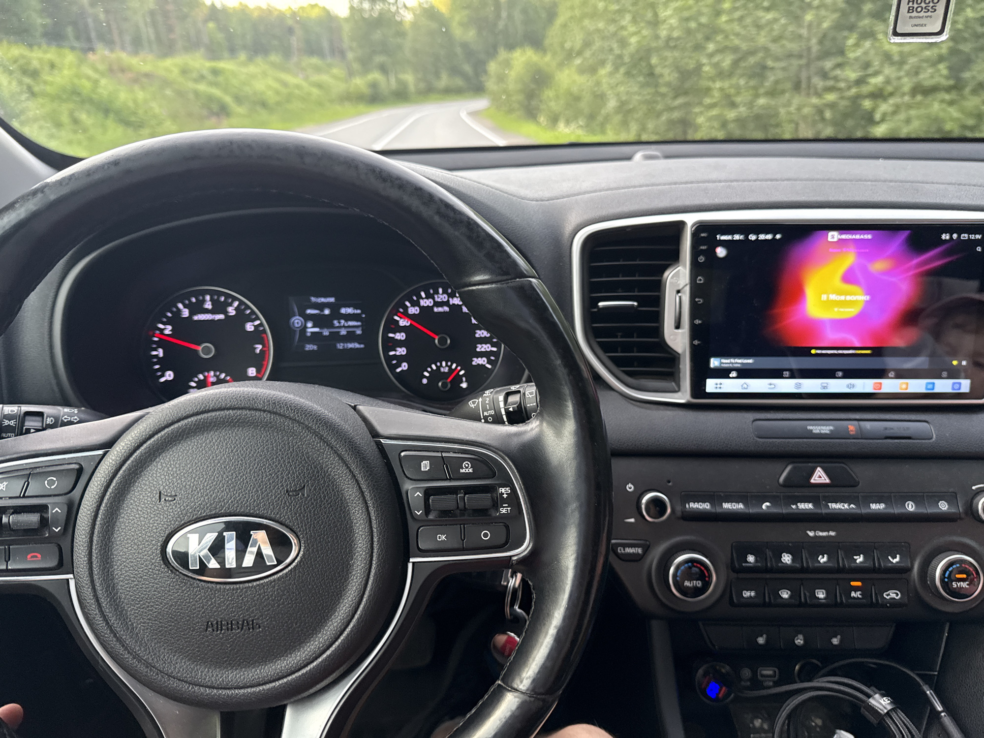 This media file captures the interior of a car, likely a Kia Sportage based on the steering wheel logo, as it drives along a road. The perspective is from the driver's seat, showing the steering wheel, dashboard, and infotainment screen.
The primary subjects are the car's interior elements, with the steering wheel and dashboard being most prominent. The infotainment screen displays album art with the text "MEGIABASS" and "11 Моя волна" (which translates to "My Wave 11" in Russian). A Hugo Boss air freshener is visible hanging from the rearview mirror.
The scene is set on a road surrounded by lush green trees, suggesting a rural or suburban location. The lighting indicates it is either dawn or dusk, with soft, warm light filtering through the trees. The weather appears to be pleasant, with no visible rain or harsh sunlight.
The activity taking place is driving. The speedometer needle indicates a speed of around 80 km/h. The overall mood is calm and relaxed, typical of a leisurely drive. Text visible includes car controls like "AUTO," "CLIMATE," "RADIO," "MEDIA," "SEEK," "TRACK," "MAP," "OFF," "A/C," and "SYNC." The dashboard also shows the odometer reading "121949" and "496 km," along with the outside temperature "20°C." The presence of Russian text on the infotainment screen strongly supports the provided location context of Kondopoga, Russia.