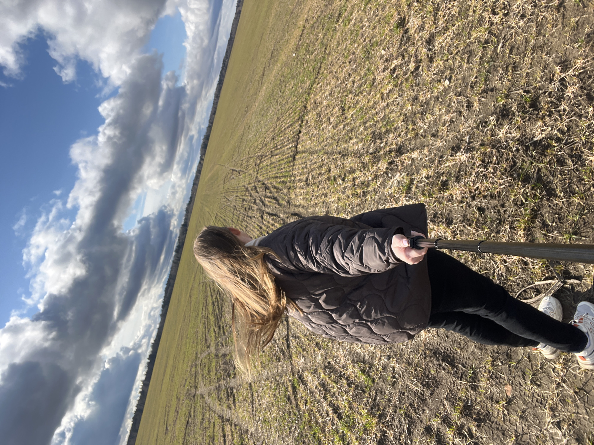 The photograph depicts a person standing in a field. The field appears to be agricultural land, with rows visible in the soil. The person is holding a selfie stick. They are wearing a brown quilted jacket, black pants, and white sneakers. The person has long blond hair. In the background, there is a view of the sky with clouds and blue patches of sky. The photograph is taken at an angle. The city of Ямуга, Russia is not visually verifiable.