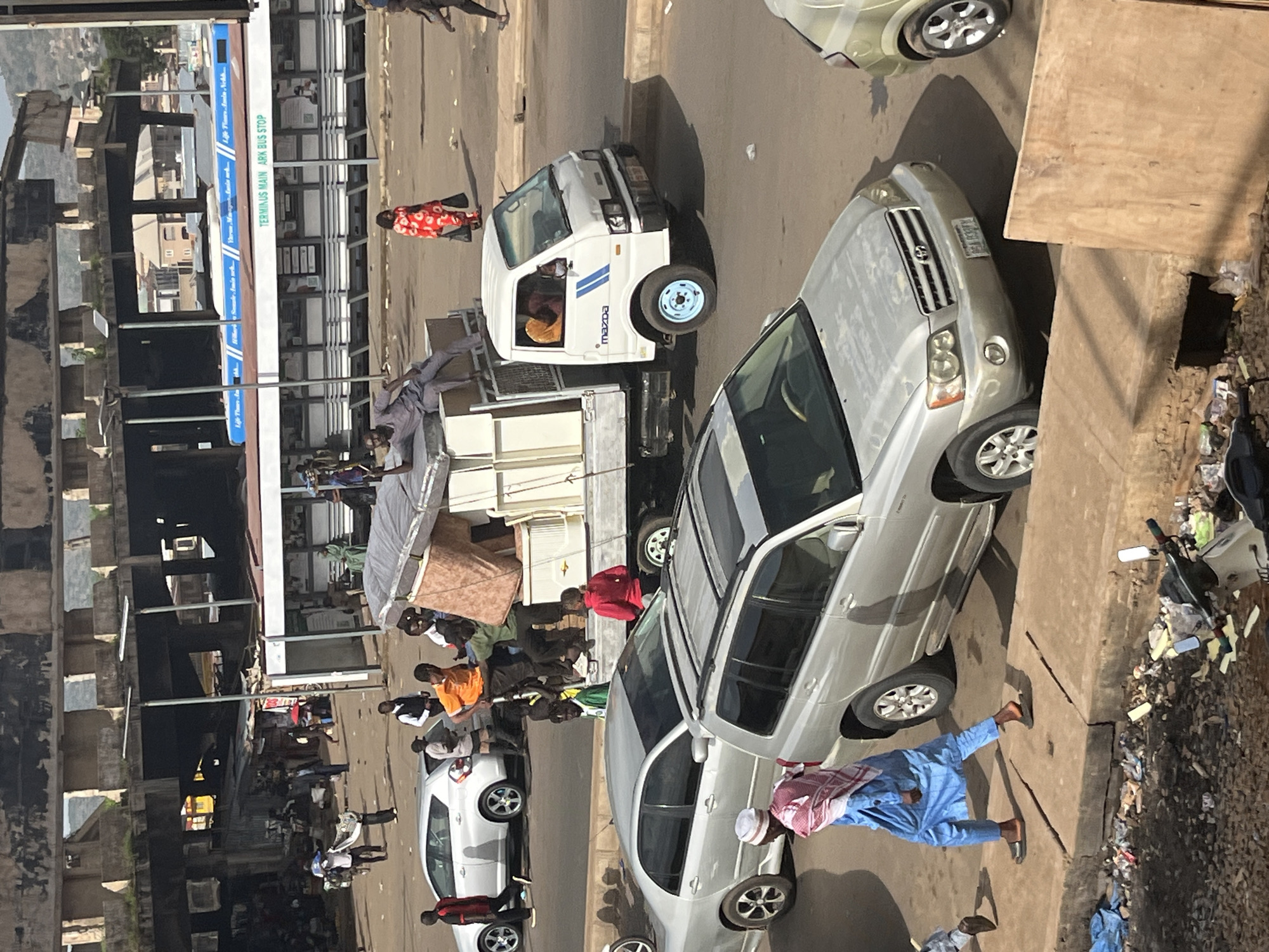 In Jos, Nigeria, a busy street scene unfolds under an overpass. A white Mazda truck, loaded with furniture, is parked on the road, with several people actively engaged in loading or securing the cargo. A silver sedan is parked behind the truck, and a silver SUV is in the foreground, partially obscuring another vehicle. A man in a blue outfit and a headscarf walks on the sidewalk, which is lined with debris and trash. The background reveals a market area with stalls and a bus stop, indicating a commercial and transportation hub.