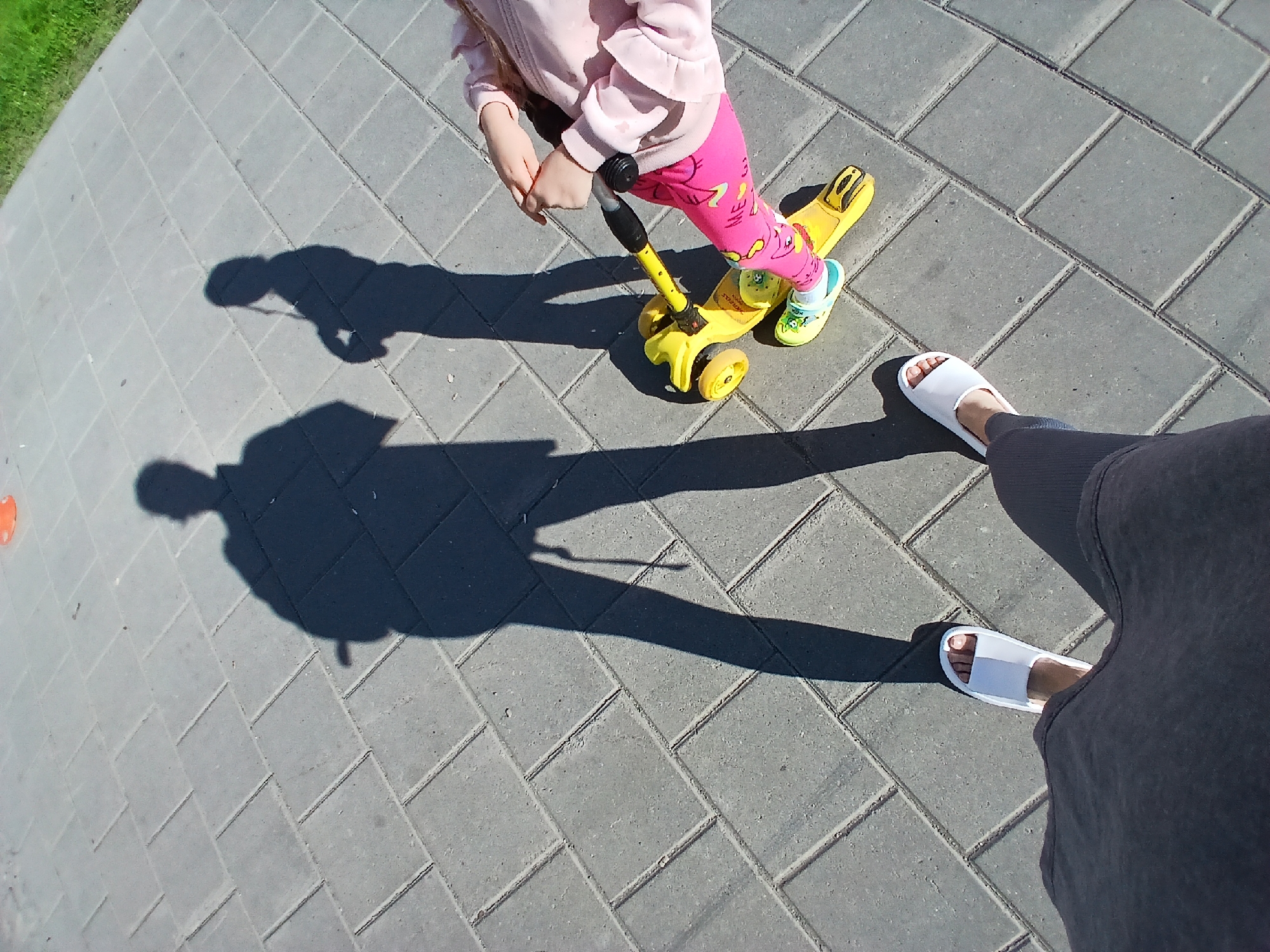 The image shows a person's shadow cast on a paved walkway in Yekaterinburg, Russia. The shadow is that of a person holding a scooter. Another, smaller shadow, presumably of a child, is also visible. The child is on a yellow scooter, wearing a pink jacket and bright pink leggings. The pavement is made of grey rectangular pavers arranged in a diamond pattern. The sun is shining brightly, creating sharp shadows. The edge of a grassy area is visible in the background. The person casting the larger shadow is wearing dark clothing and white slides. The child's feet are clad in yellow slides with a blue and white sock.