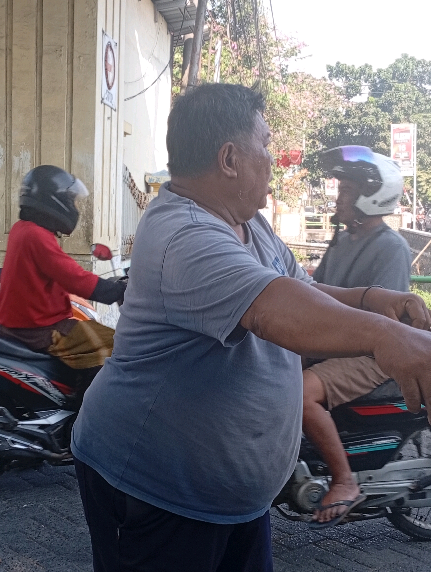 The image captures a scene on a street in Jogosari, Indonesia. In the foreground, a man with a large build is riding a motorcycle, facing towards the right side of the frame. He is wearing a gray t-shirt and dark pants. His right arm is extended, resting on the handlebars of his motorcycle, and his left arm is also visible. The motorcycle appears to be black and silver.
To the left of the man, another person is riding a motorcycle, seen from the side. This individual is wearing a red long-sleeved shirt and a black helmet with a visor. They are also wearing what appear to be brown or tan pants. Their motorcycle is black and orange.
In the background, another person is visible on a motorcycle, wearing a white helmet with a blue visor and a gray t-shirt. This individual is also facing to the right.
The environment suggests an outdoor, urban setting. There is a building with a light-colored, vertically grooved facade on the left side of the image. A white sign with a red circle and a diagonal line is attached to this building. Trees with pink blossoms are visible in the background, along with other foliage and overhead wires. The ground is paved. The overall lighting suggests a daytime scene.