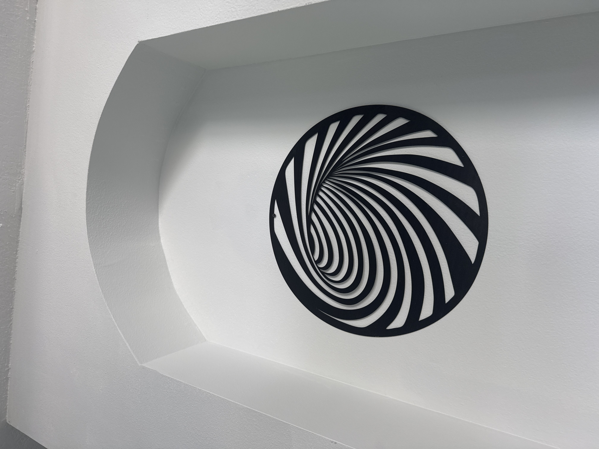 The image shows a black, circular, optical illusion art piece mounted on a white wall. The art piece features a pattern of black and white stripes that create a sense of depth, resembling a vortex or tunnel. The wall itself is within a recessed architectural feature, possibly a niche or alcove, with curved archways. The lighting in the scene casts soft shadows, highlighting the texture of the wall. The location is Viera, United States of America.