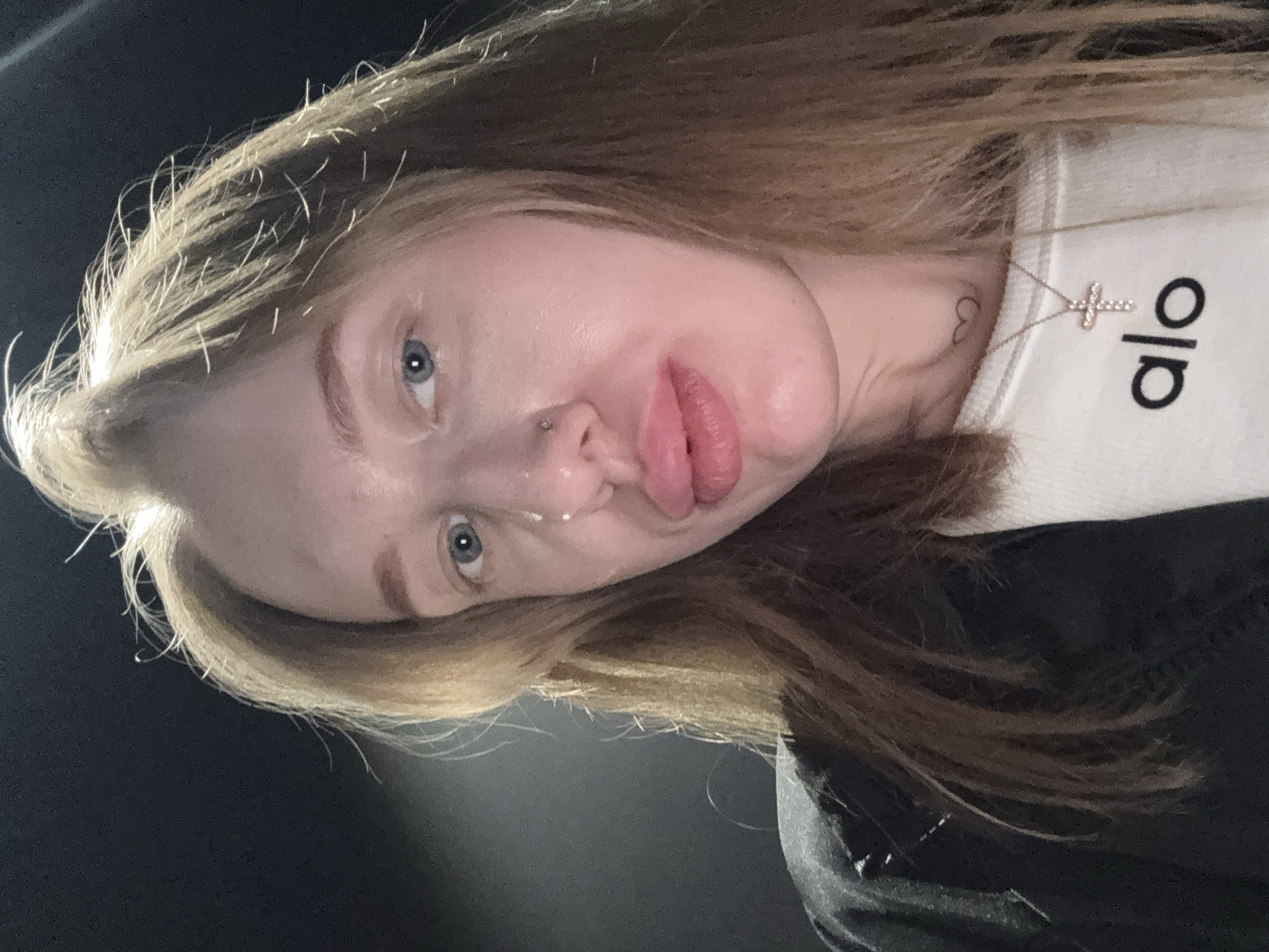 A close-up, slightly rotated image of a fair-skinned person with light brown hair. They have blue eyes, a nose piercing, and are making a pouting expression. They are wearing a white shirt with the word "alo" printed in black on it, and a silver necklace with a cross pendant. A small heart tattoo is visible on their neck. The background is a dark, out-of-focus gray/black. The location is not discernible from the image.