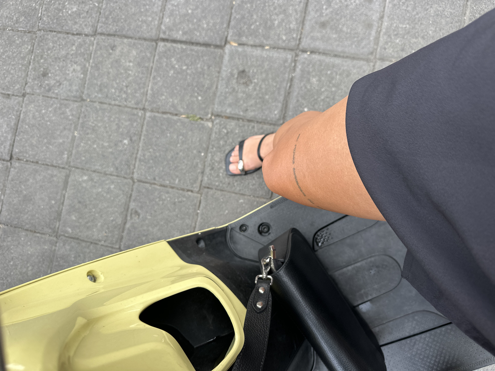 A high-angle shot shows a person seated on a scooter, possibly in Mengwi, Indonesia, but no specific landmark is visible to confirm the location. The individual wears a black dress and black sandals with a decorative ring on the big toe. A black leather purse hangs on the scooter. The scooter is primarily yellow. The ground consists of grey paving stones. The individual's leg features a tattoo.
