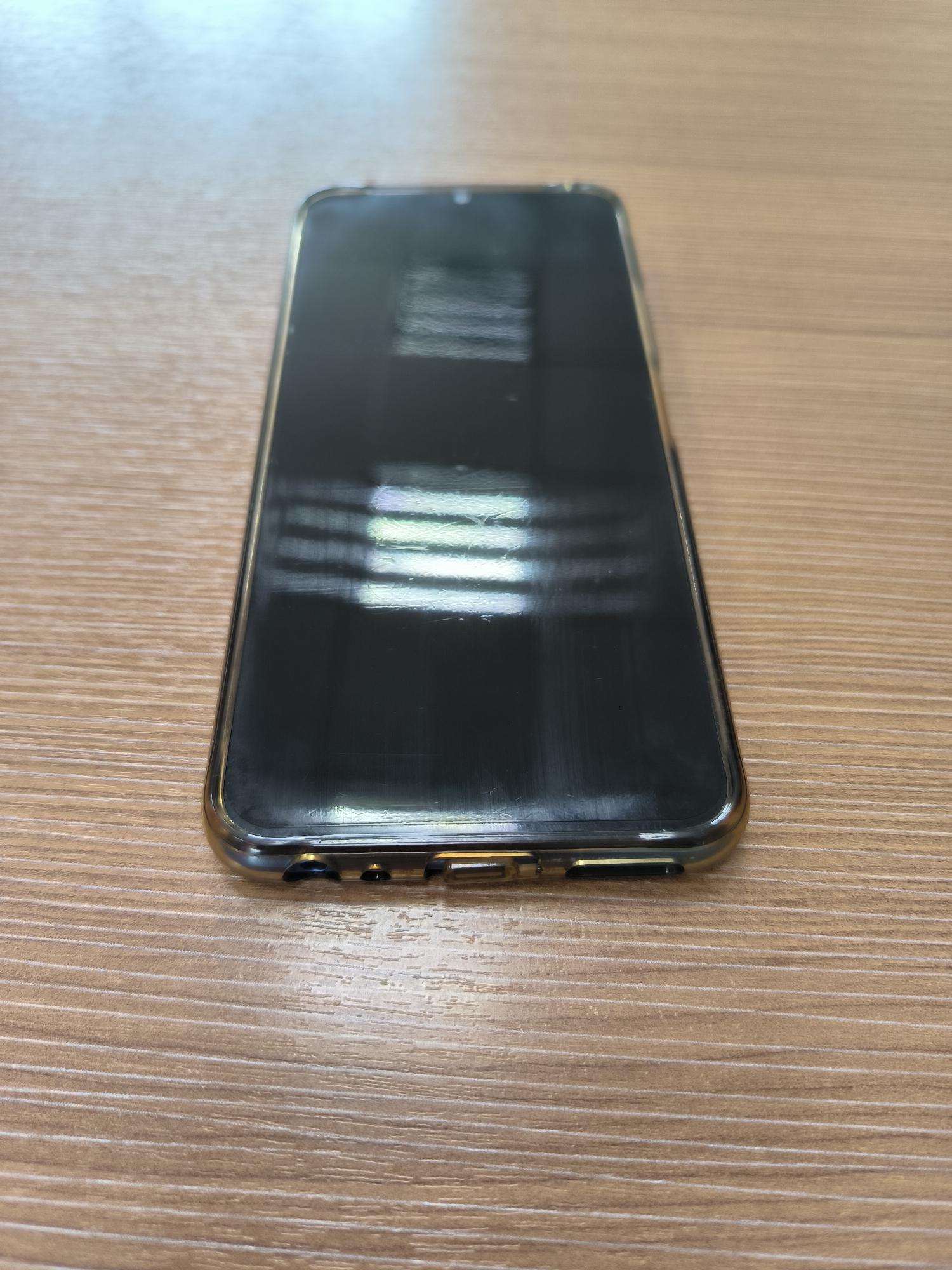 A smartphone with a clear case rests on a wooden desk. The phone's screen reflects light, showing horizontal lines. The desk has a visible wood grain. The scene is indoors, likely a home or office environment. The image is taken from a slightly elevated angle, looking down at the phone. There are no people or animals visible in the frame. The lighting suggests it is daytime, possibly from a window or overhead light source. No specific location in Yuzhno-Sakhalinsk, Russia can be determined from the image.