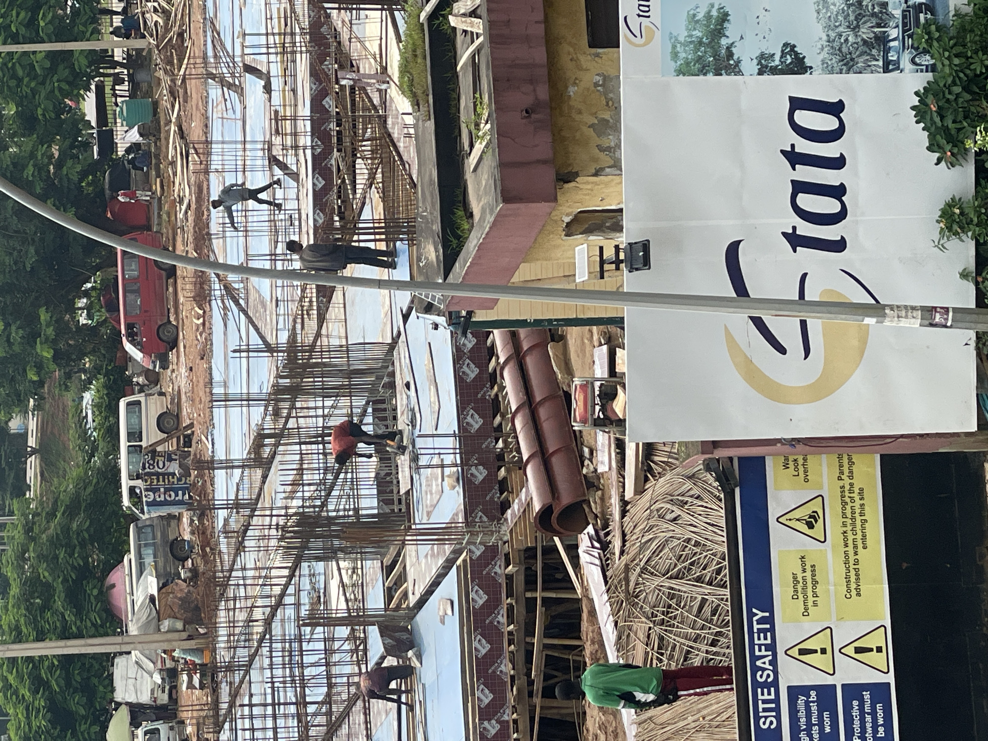 This image depicts a construction site in Abuja, Nigeria, likely in the Garki district, characterized by ongoing building activities. The scene includes multiple construction workers actively engaged in their tasks. One worker, wearing a red shirt, is seen operating a power tool on a concrete platform reinforced with rebar. Another worker, in a grey shirt, stands on the same platform, seemingly overseeing the work. Further in the background, a worker in a red shirt is also visible, and another in a grey jacket is standing on a higher level of the structure. In the foreground, a person wearing a green shirt and red pants stands facing away from the camera, observing the construction.
The environment shows a large, multi-story building under construction with extensive scaffolding and concrete work. The ground is a mixture of red earth and scattered construction debris, including bundles of reedy material, cylindrical concrete molds, and various metal components. Several vehicles are present, including a red minivan and a white van, suggesting logistical support for the site. In the distance, lush green trees indicate an urban setting with established vegetation.
A prominent white billboard with the word "tata" in blue lettering is visible in the foreground, partially obscuring the construction activity. Beside it, a "SITE SAFETY" sign lists several cautionary notes, including "High visibility jackets must be worn," "Protective footwear must be worn," and "Danger Demolition work in progress," accompanied by warning symbols. The overall impression is one of an active construction zone with a focus on safety regulations.