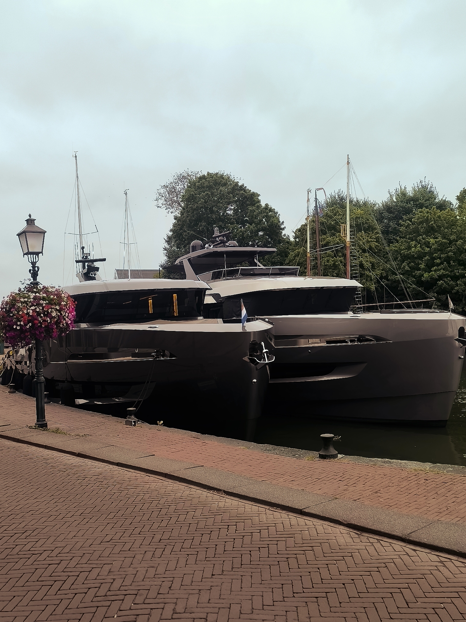 The image shows a cloudy day in Hoorn, Netherlands, with two modern yachts docked along a brick-paved waterfront. A lamppost adorned with a hanging basket of colorful flowers stands on the left side of the frame. The yachts are sleek and dark, with large windows and multiple levels. One yacht appears to be a catamaran. Trees line the background, suggesting a park or natural area behind the quay. The water is dark and still. The scene conveys a sense of quiet elegance.