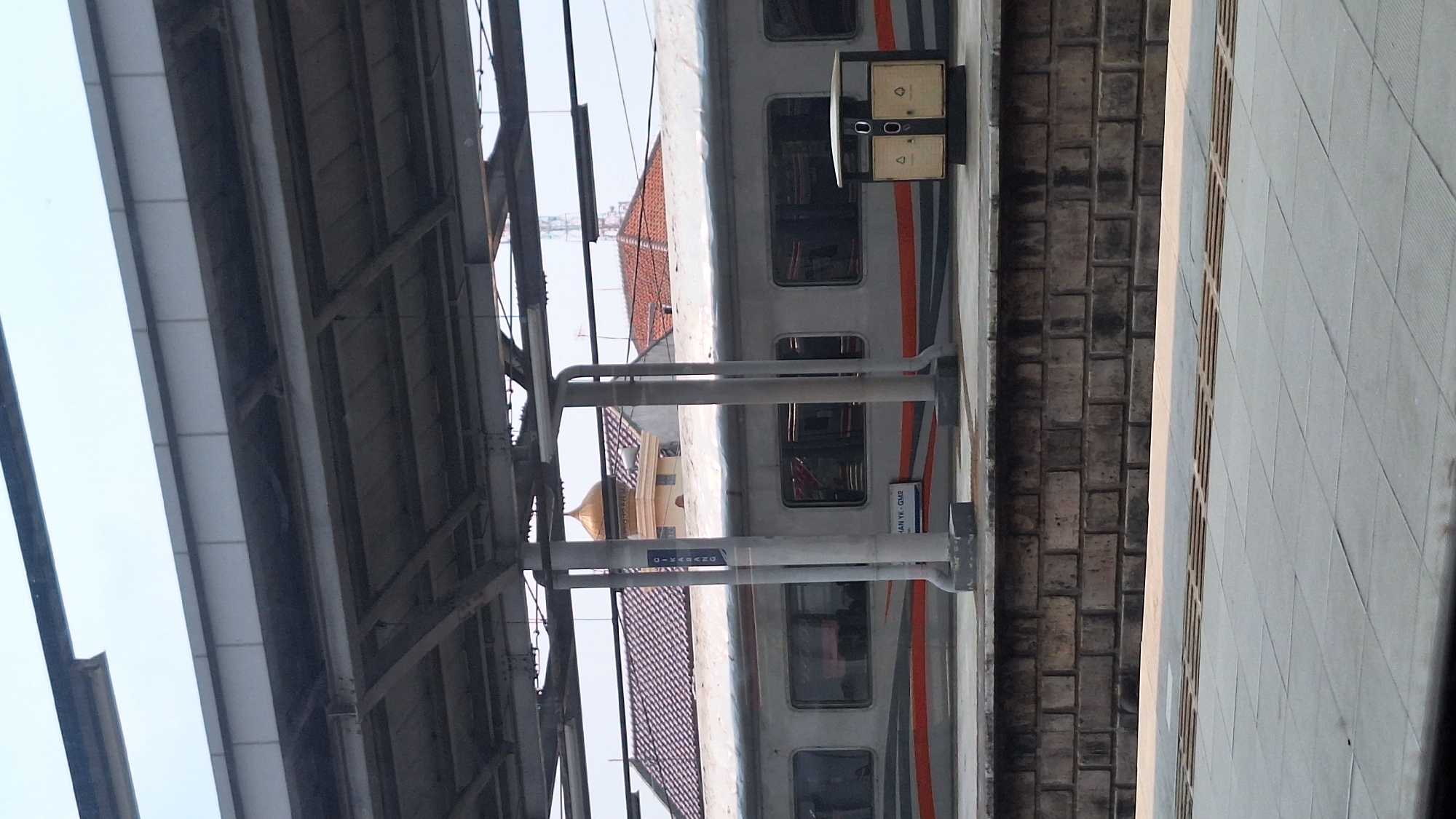 The image shows a train stopped at a station. The station appears to have an overhead shelter structure with metal beams and a corrugated roof. Adjacent to the train is a platform area with textured concrete and a brick wall separating the tracks. In the background, building roofs with red tiles and a golden dome are visible, and a radio transmission tower. A sign on a support column reads "DIKSANO" indicating the specific location may be Cikarang, Indonesia. The train itself is primarily white with orange and grey stripes.
