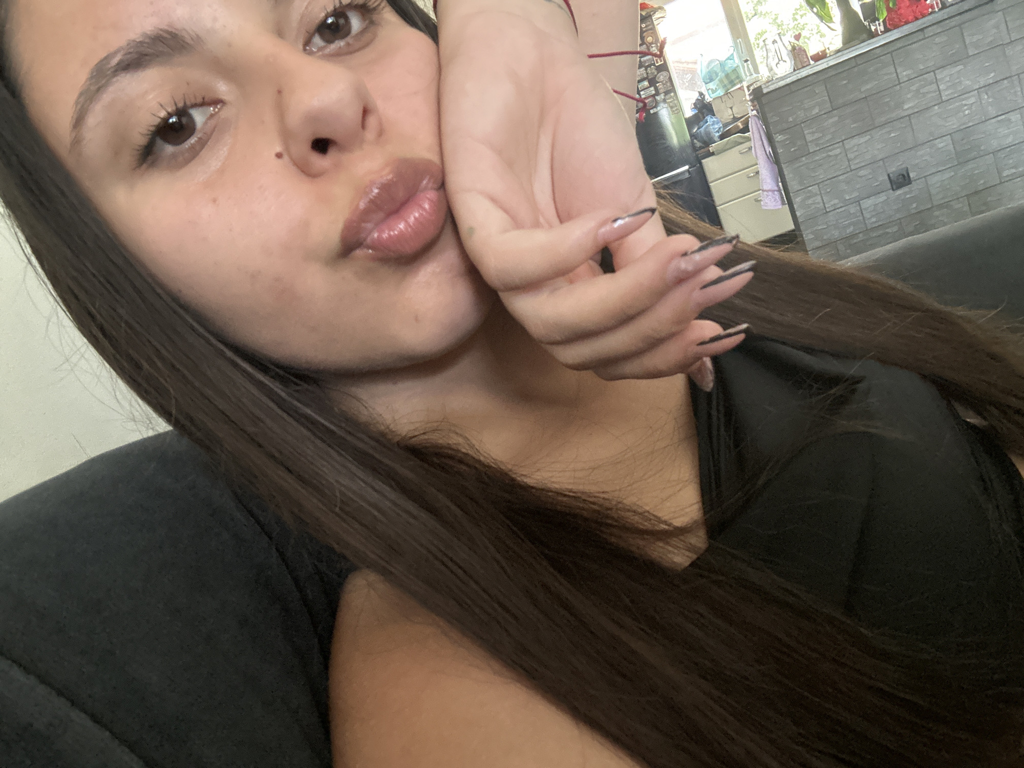 A close-up, slightly tilted selfie captures a young woman from the chest up, showcasing her face, hair, and a hand. Her long, dark brown hair cascades over her shoulders and chest. She has dark brown eyes and appears to be wearing light makeup, with full, glossy, light-colored lips pursed in a "kiss" or "pout" expression. Her left hand, adorned with long, dark-tipped (likely black or dark grey) nails, is resting against her face, partially obscuring her left cheek. A thin red string bracelet is visible on her wrist. She is wearing a dark, possibly black, casual top or t-shirt.
The background is a domestic or casual indoor setting, illuminated by natural daylight streaming through a window on the right. Visible elements include a section of a grey, textured wall, possibly tiled or stone, with a dark electrical outlet. A white cabinet with drawers is partly visible, along with some green plants and red flowers. Further in the background, a dark punching bag can be faintly seen. The overall scene suggests a relaxed atmosphere, possibly in a living room, bedroom, or a casual shop. No visible text is present in the image.