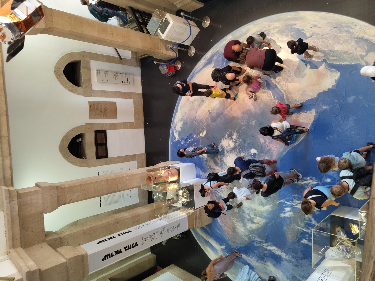 This is an elevated, wide-angle shot of an indoor exhibit, likely in a museum, featuring a large, floor-mounted map of the Earth. The map is a prominent central element, with many people of various ages interacting with it. The overall environment suggests a public space, possibly a science or history museum.
Several individuals are depicted standing on or around the Earth map. Some are pointing, while others are observing or walking. There are adults and children present, indicating a family-friendly atmosphere. Notable figures include a person in a blue backpack descending a staircase on the right side of the frame, and several people gathered around the Earth map in the center, some appearing to be explaining or demonstrating something. A satellite model is visible suspended from the ceiling in the upper right corner.
The architecture suggests an older building, with stone pillars and arched doorways visible. Banners with text in what appears to be Hebrew are hanging from the pillars on the left. Display cases containing artifacts are positioned around the exhibit space, hinting at the museum's collection. The lighting appears to be artificial, typical of an indoor exhibition. The floor is dark, contrasting with the vibrant blue and white of the Earth map.
The presence of Hebrew text on the banners, coupled with the general style of the architecture and the overall theme of the exhibit (the Earth and possibly space exploration, given the satellite), strongly suggests this location is in Israel. Considering the prompt's mention of Haifa, Israel, and the historical architectural style often found in museums there, it's plausible this scene is within a museum in that city.