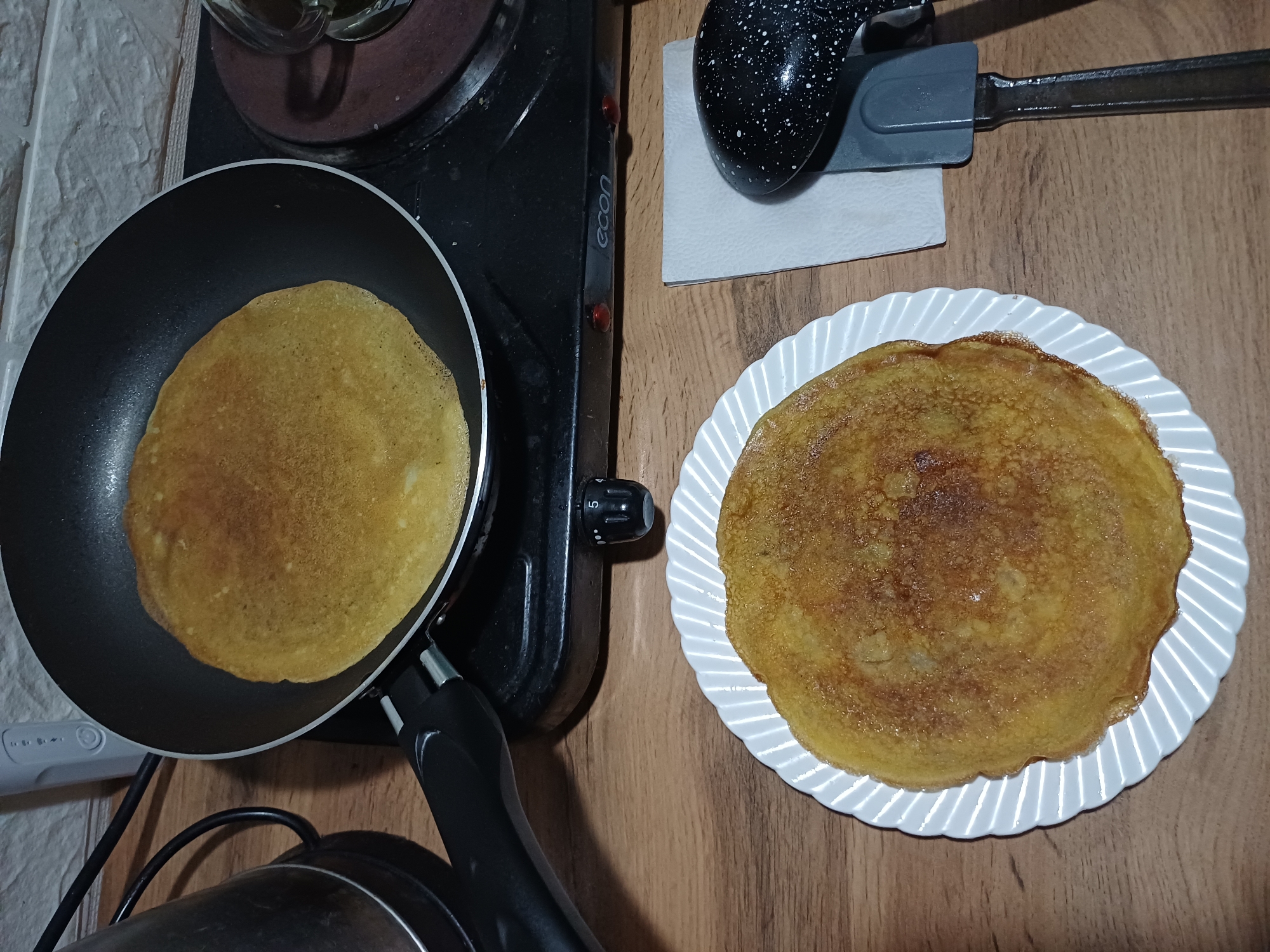 In a domestic kitchen, a pancake is being cooked in a black frying pan on a portable electric stovetop. The pancake is golden brown and appears to be partially cooked. Next to the stovetop, a white plate with a finished pancake sits on a wooden countertop. A spatula with a black speckled handle rests on a white paper napkin. The scene is dimly lit, suggesting it may be evening or early morning. There is no indication of the location being in the city of Sochi, Russia.