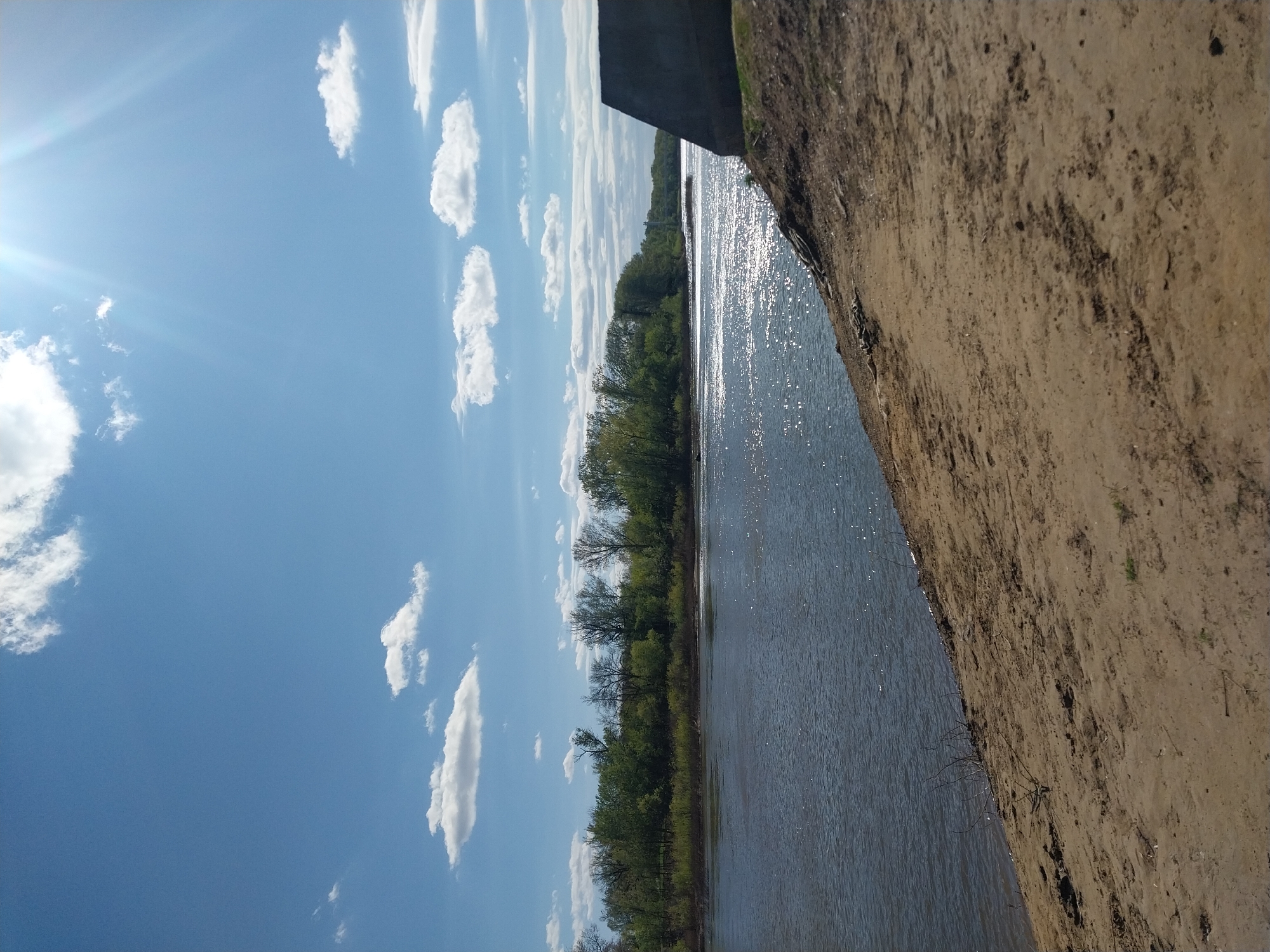 The image shows a landscape with a river running vertically through the center. On the left of the river, there is a blue sky with scattered white clouds. On the far side of the river, there is a line of green trees. To the right of the river is a bank covered in dirt and sparse vegetation. The photograph appears to have been taken next to a dirt bank, possibly a road or a large levee.
While it shows a river and landscape, without additional information, it is impossible to confirm that the photo was taken in Orenburg, Russia.