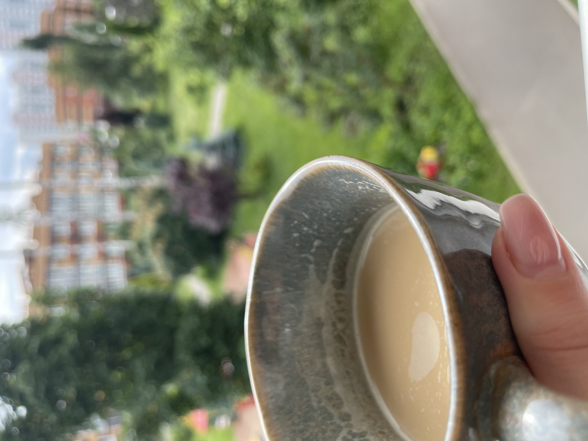A close-up shot focuses on a ceramic mug filled with a light brown beverage, held by a hand with pink-painted fingernails. The mug has a textured grey-brown glaze. In the background, a blurred scene depicts greenery, trees, and multi-story buildings. The setting appears to be an urban residential area, possibly in the city of Kommunarka, Russia, given the presence of apartment buildings and manicured green spaces. The lighting suggests it is daytime, with overcast skies contributing to the soft illumination.